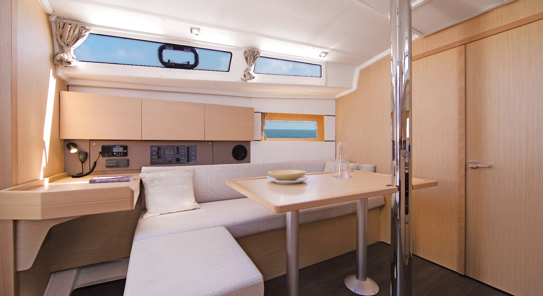 Interior view of a Beneteau Oceanis 38.1 yacht cabin with wooden walls, a small dining table, and a cushioned bench seat. There are windows showing the sea outside, with curtains and built-in storage units.
