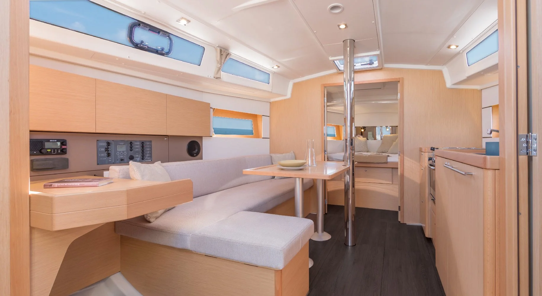 Interior of a Beneteau Oceanis 38.1 yacht cabin with wooden cabinetry, a small dining area with a beige cushioned bench and table, a mirrored door leading to a sleeping area with bed, and windows letting in natural light.