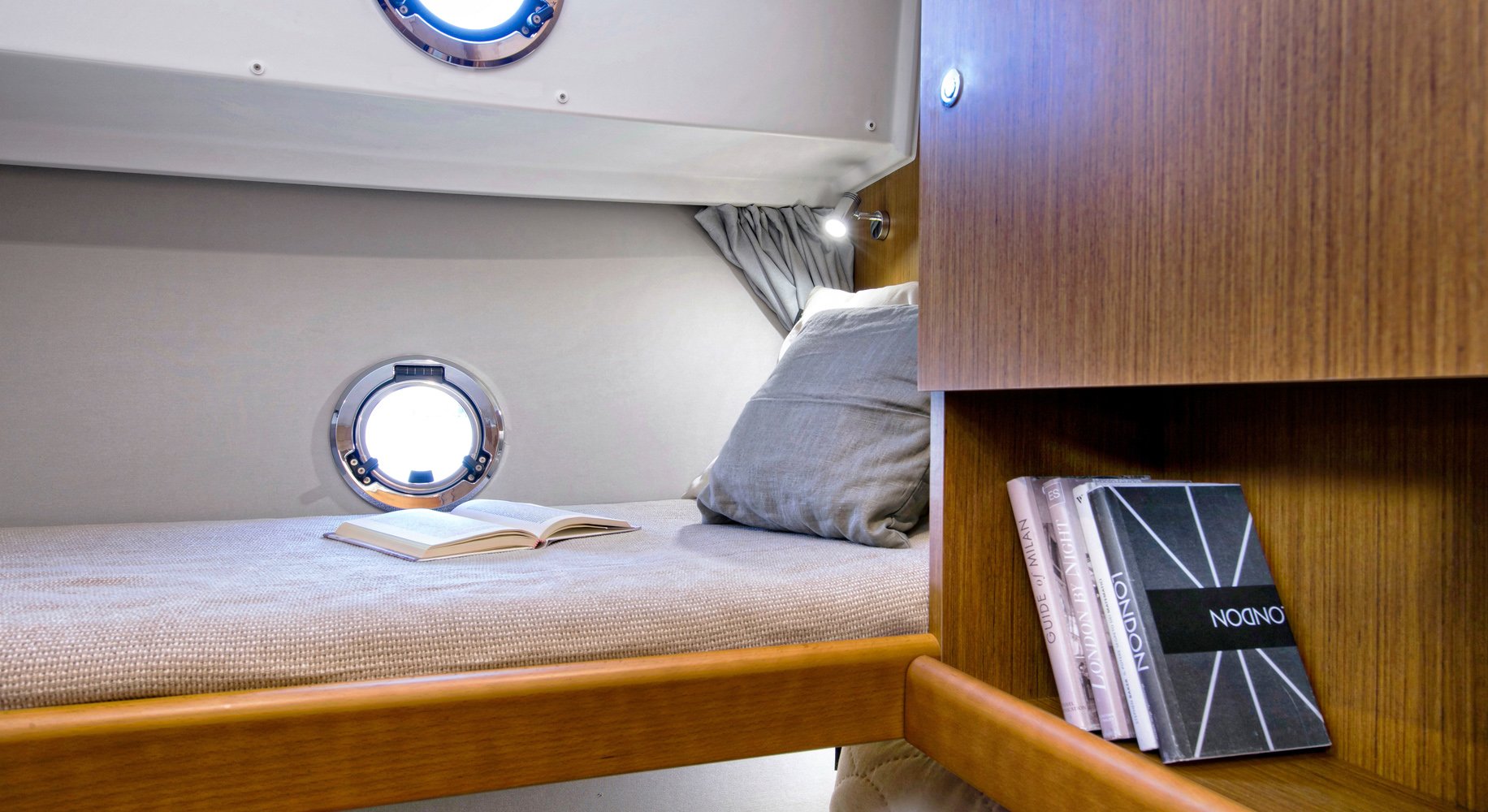 A cozy bedroom interior of a Beneteau Swift Trawler 35 with porthole windows on the wall, a bed with a gray pillow and beige bedding, an open book, and a bookshelf with notebooks and books.