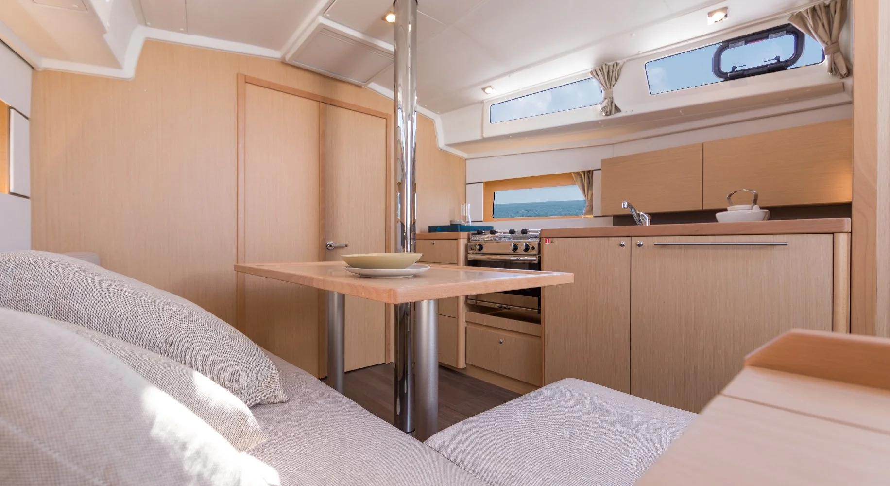 Interior of a Beneteau Oceanis 38.1 yacht cabin featuring a kitchenette with light wood cabinets, a small stove, and windows with curtains. There is a beige sofa with pillows and a small wooden table with a bowl and plate.