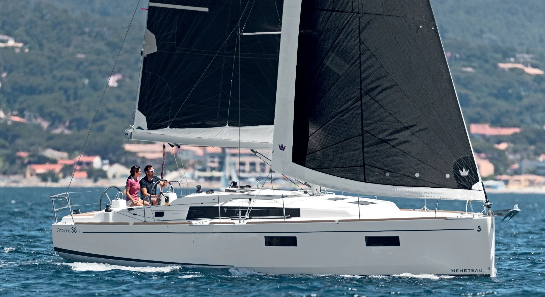 A Beneteau Oceanis 38.1 sailboat with black sails on the water, with two people on board, sailing near a coastline with buildings and greenery in the background.