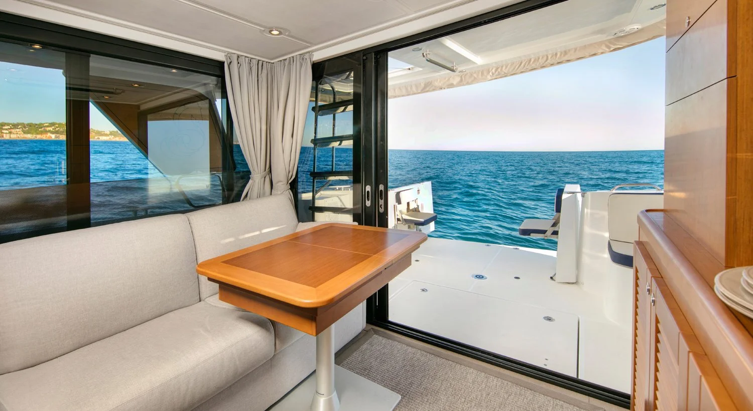 Interior of a Beneteau Swift Trawler 35 yacht with a glass sliding door open to the ocean view, featuring a beige sofa, a wooden table, and a wooden cabinet, with a deck outside and a ladder leading up to the higher deck.