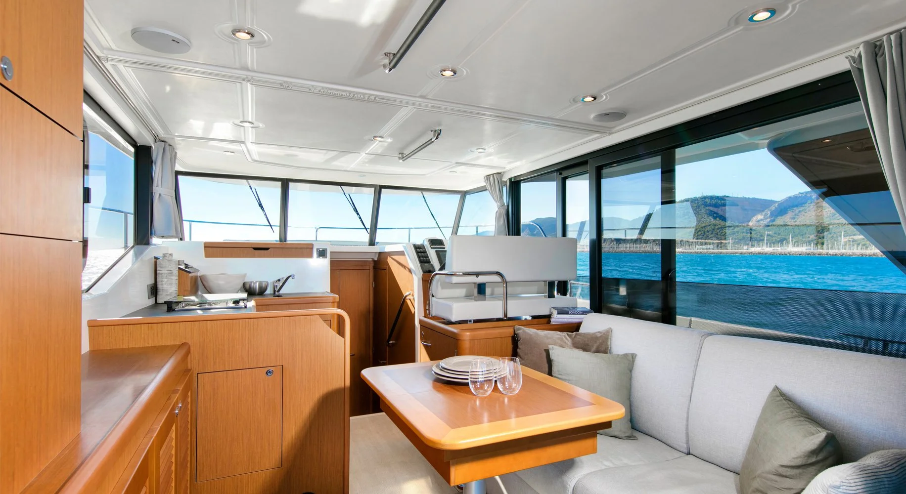 Interior of a Beneteau Swift Trawler 35 yacht with large windows showing a view of water and mountains, featuring a small kitchen area with wooden cabinets, a dining table with plates and glasses, and a white sofa with pillows.