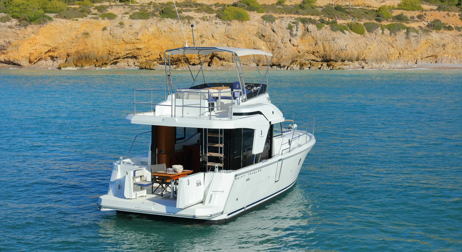 A Beneteau Swift Trawler 35 sailing in calm blue waters near a rocky coastline with sparse greenery.
