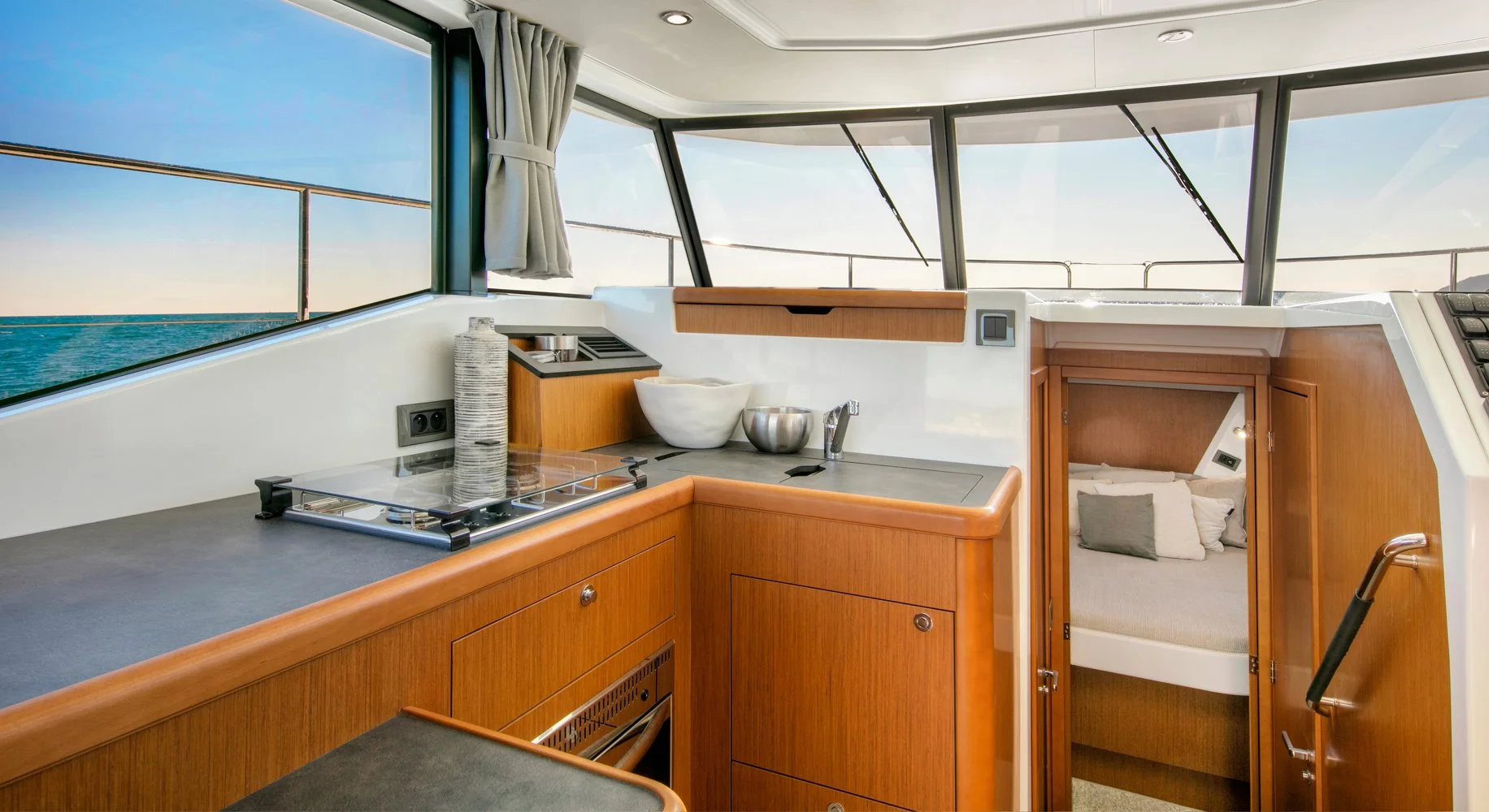 Interior of a Beneteau Swift Trawler 35 cabin with a compact kitchen area featuring a countertop, sink, and small stovetop, with a sleeping nook visible through a small door, and large windows showing a view of the water and sky.