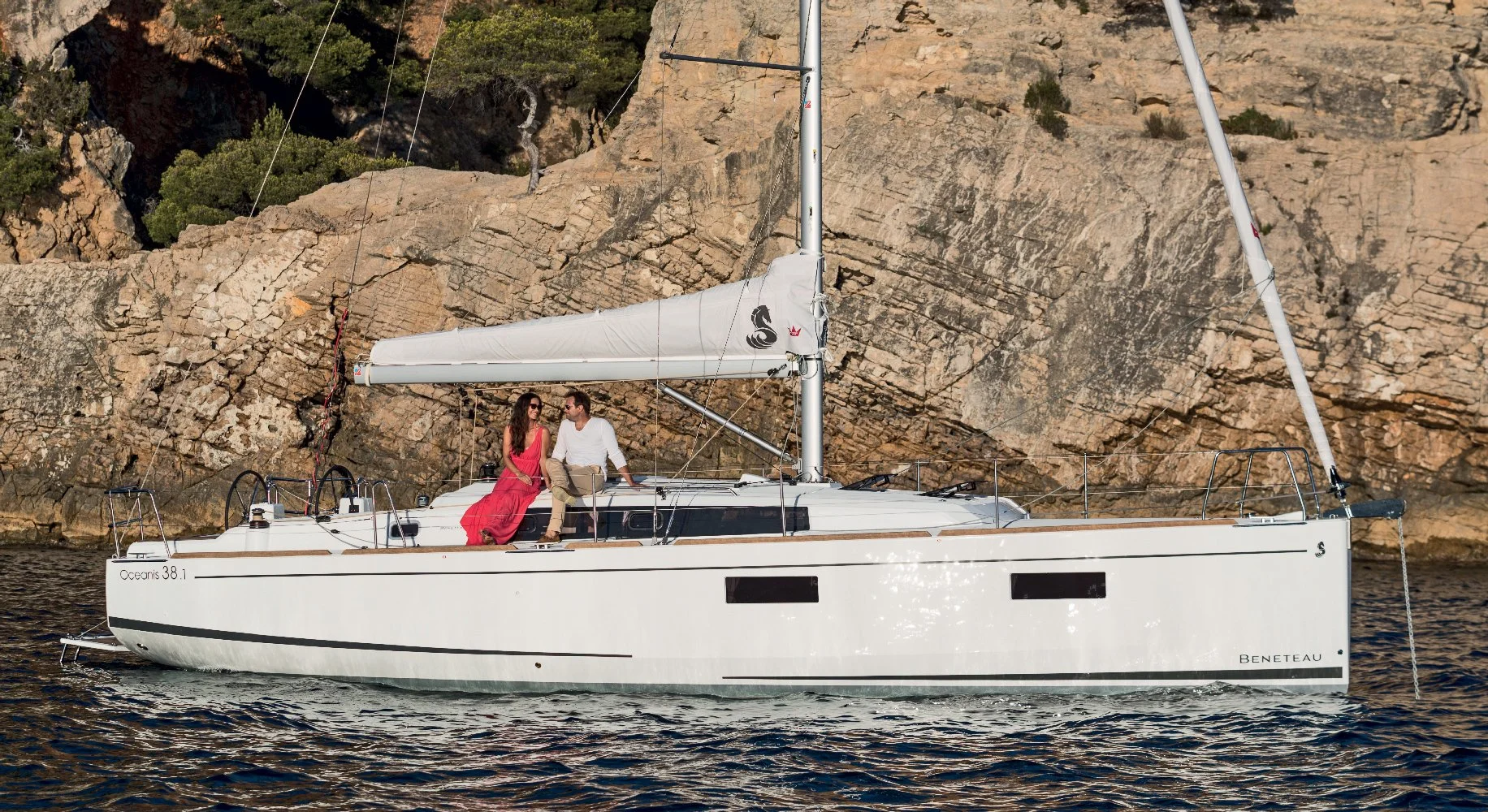 A Beneteau Oceanis 38.1 sailboat with two people sitting on the deck near a rocky shoreline with green vegetation in the background.