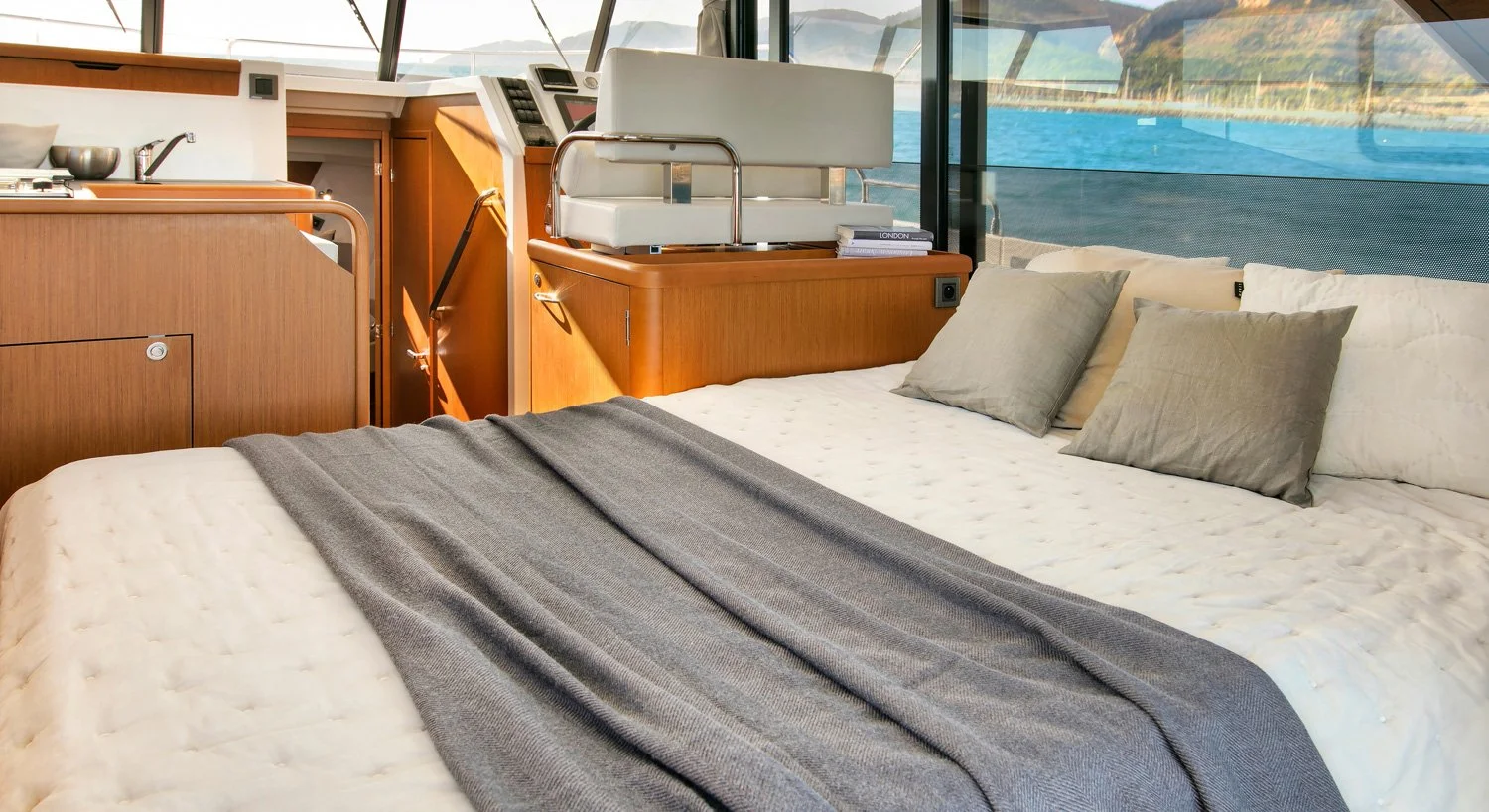 Interior of a Beneteau Swift Trawler 35 yacht showing a bed with gray pillows, a gray blanket, and large windows with a view of water and sky outside.