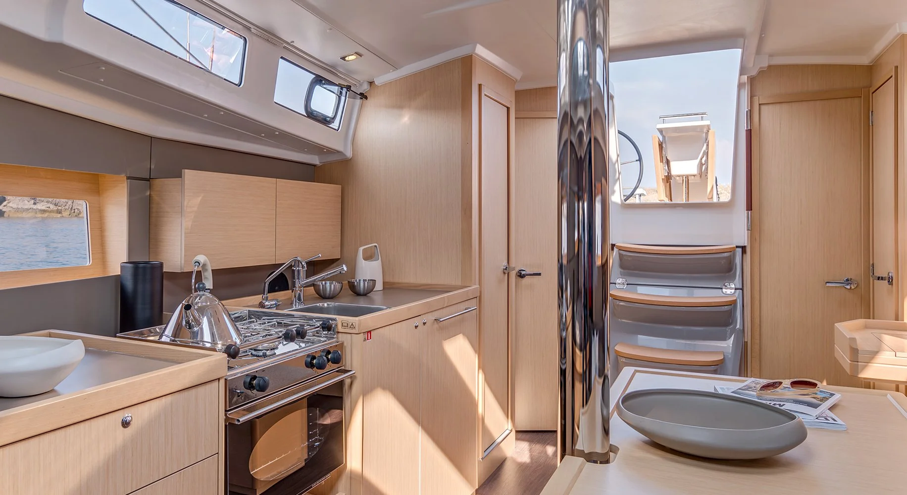Interior of a Beneteau Oceanis 38.1 kitchen with a stove, sink, cabinets, and a small window showing water outside. There are magazines and a decorative bowl on a table, with sunlight streaming in.