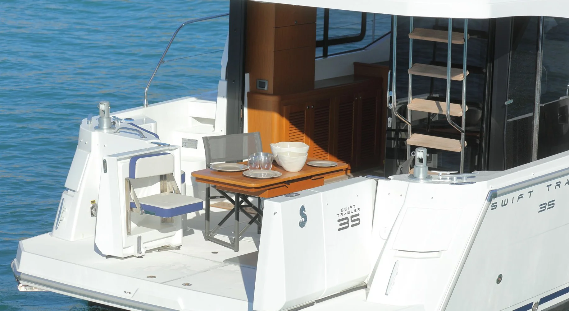 Back deck of a Beneteau Swift Trawler 35 yacht with a small folding table set with plates and bowls, a chair, and a staircase leading to the upper deck, floating on water.