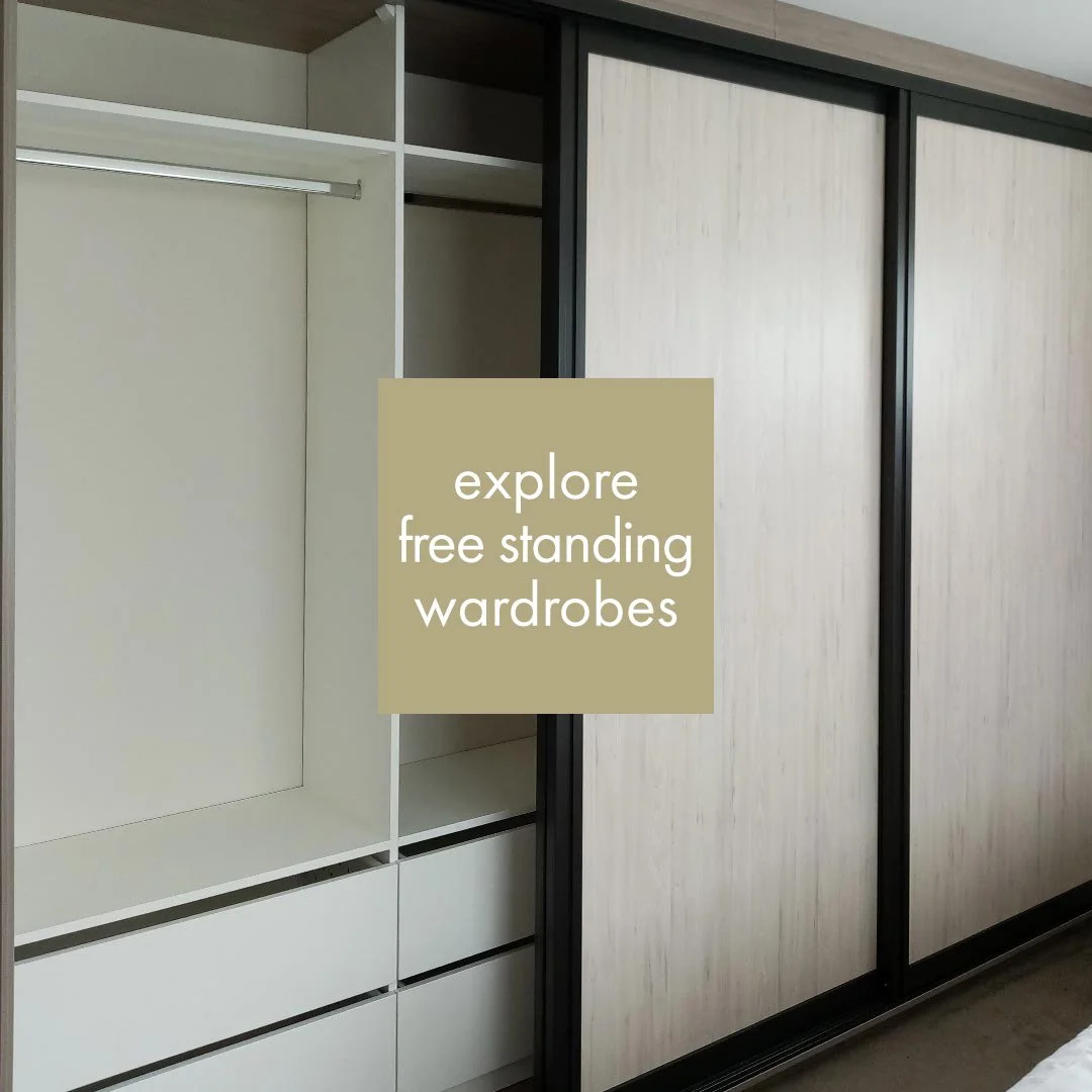An open closet with shelves and drawers next to a sliding door wardrobe with a light wood finish, with a sign saying 'explore free standing wardrobes' across it.