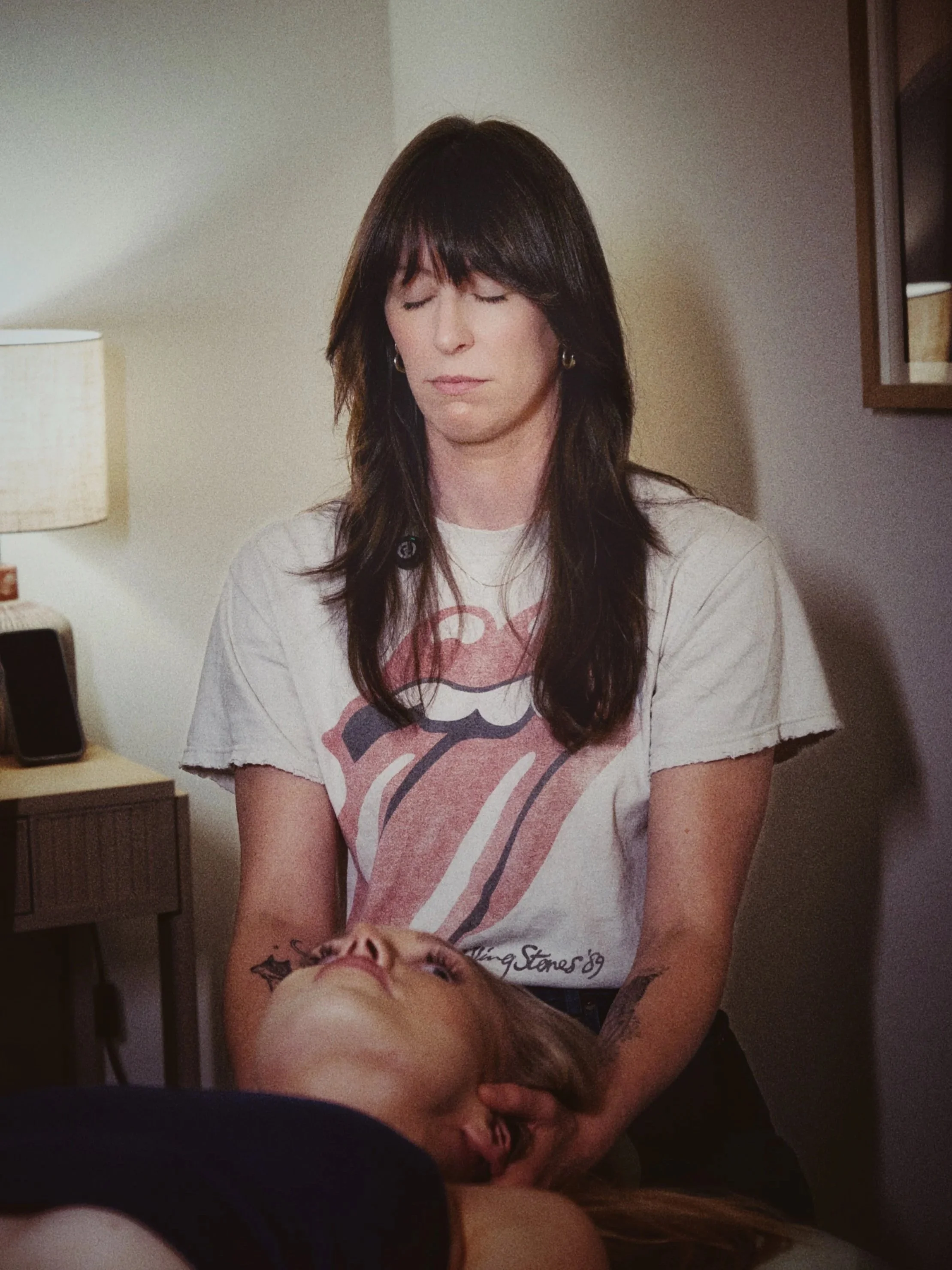 A woman with long, dark hair and a Rolling Stones t-shirt gives a massage to a woman lying on a bed or table, with her eyes closed, in a room with a lamp and some furniture.