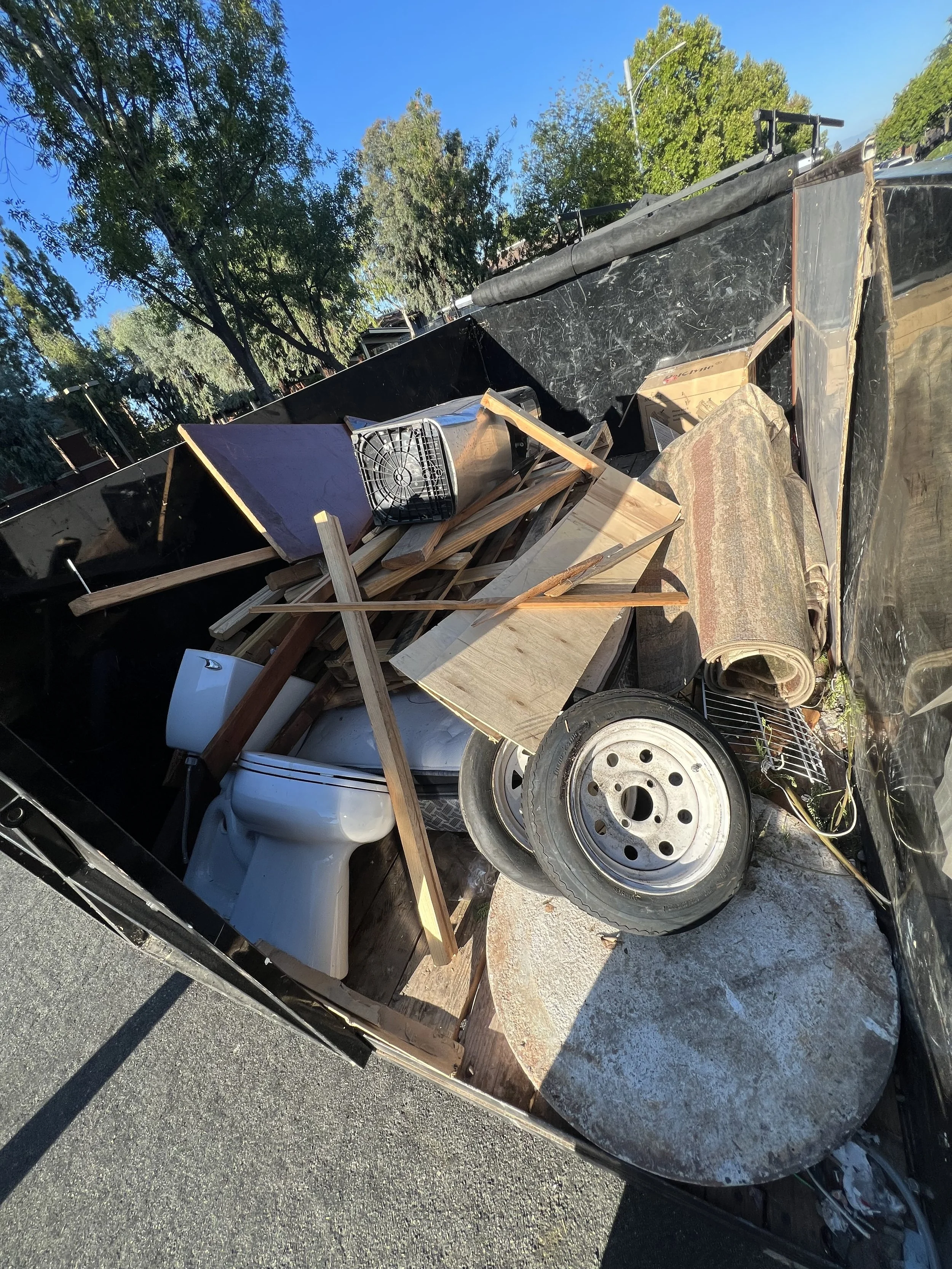 Overflowing dumpster filled with construction debris, including wooden planks, a toilet, a tire, a rolled-up carpet, and various other discarded materials, with trees and a clear blue sky in the background.