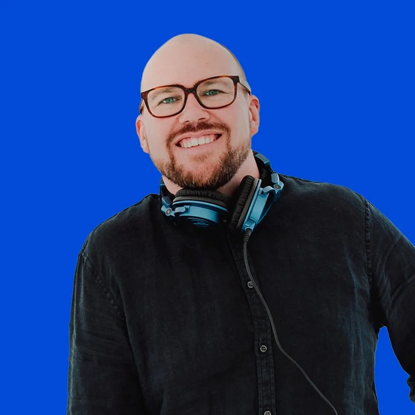 Glen James with glasses and a beard, wearing a black shirt and headphones around his neck, against a blue background.