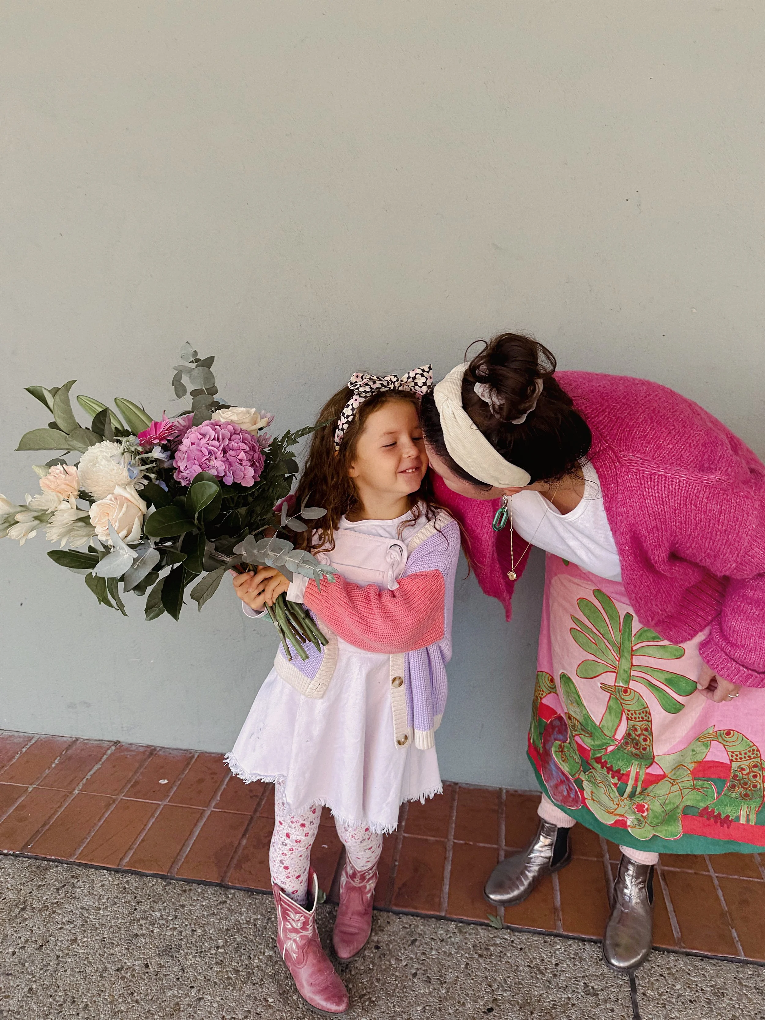 Two women sitting on a park bench, smiling, with trees and buildings in the background.