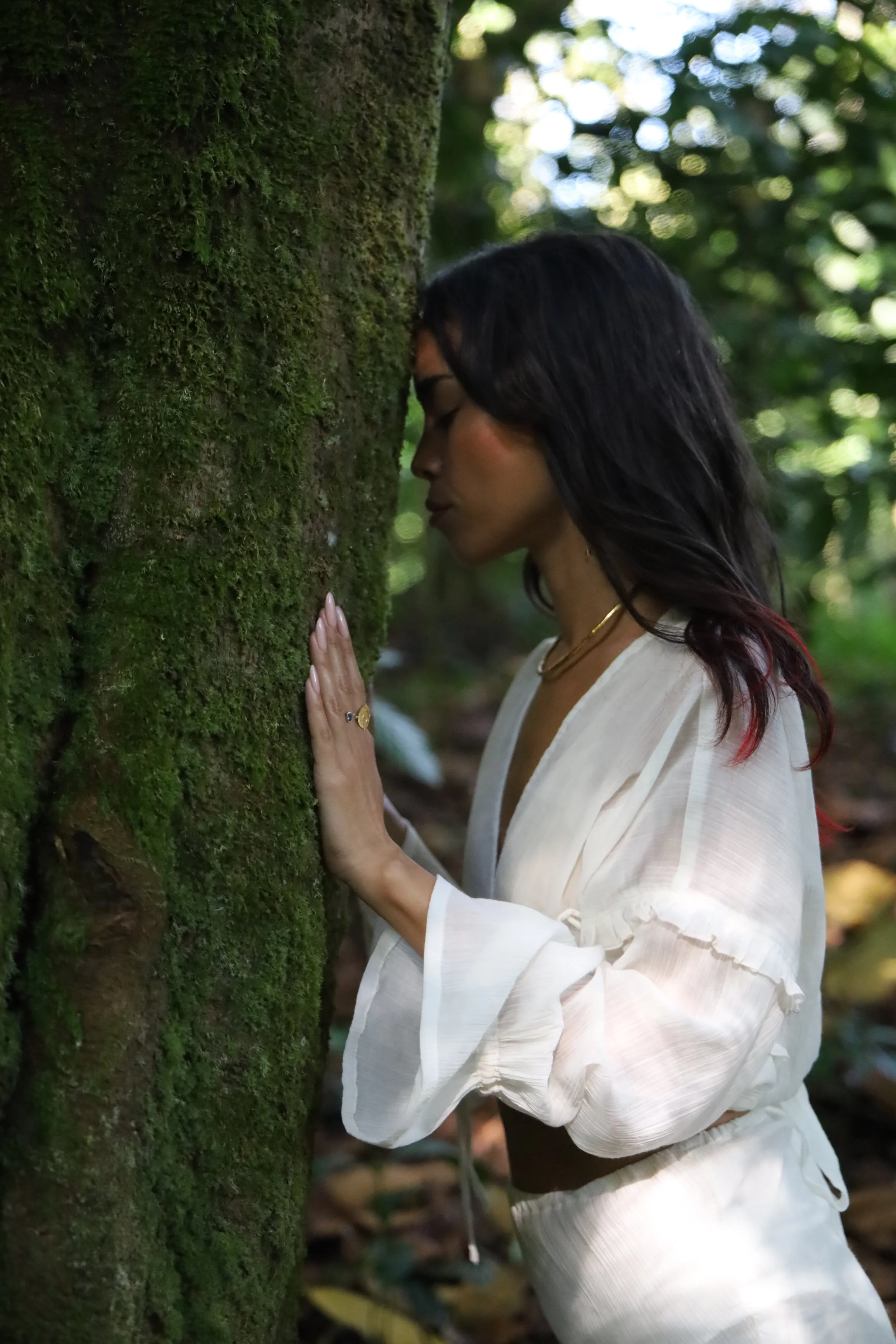 A woman with dark hair and wearing a white blouse touches a moss-covered tree with her eyes closed.