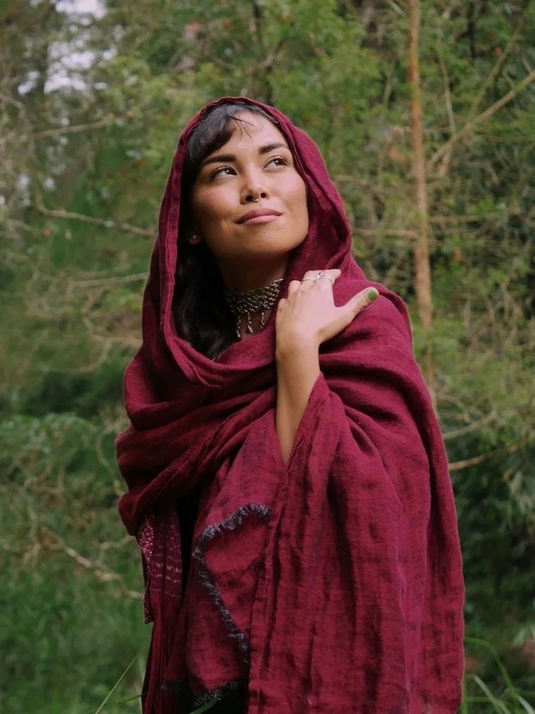A woman with dark hair and a septum piercing, wearing a maroon shawl, standing outdoors in a lush green forest, looking thoughtfully to the side.