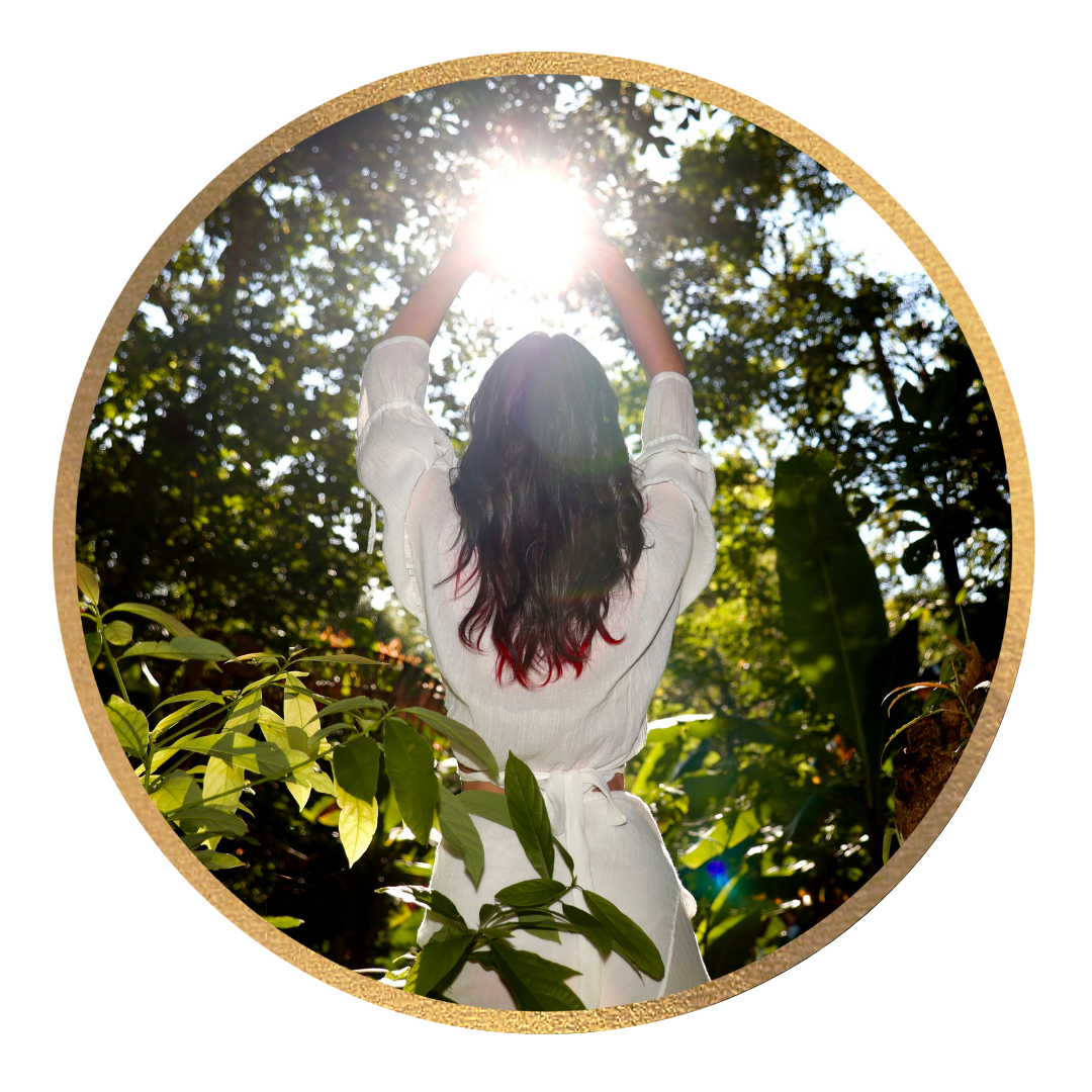 A woman with long dark hair standing outdoors in a garden or forested area, reaching up towards the bright sun with her arms raised, surrounded by green foliage and trees.