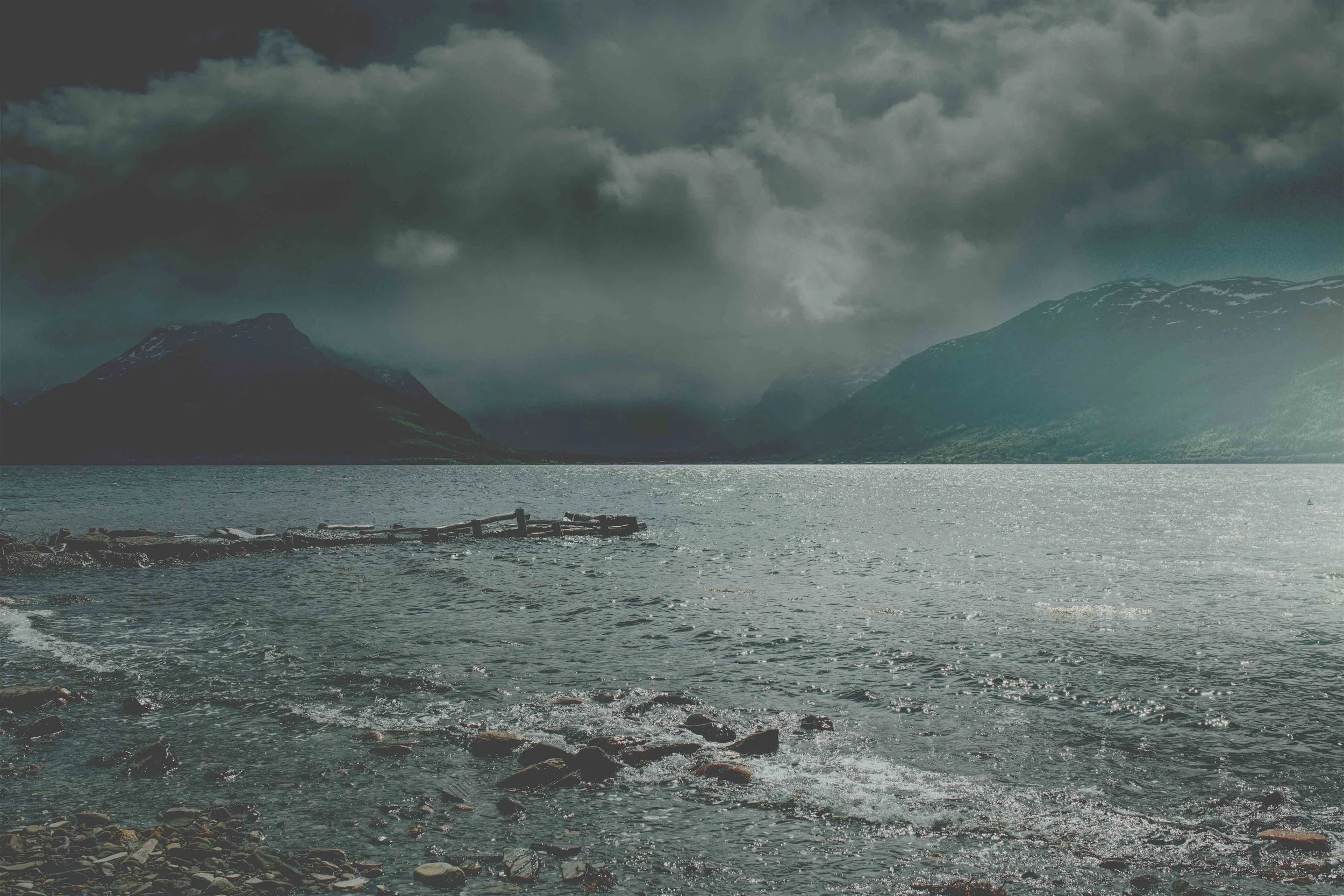 Scenic view of a mountain valley with a winding road and river, surrounded by tall mountains with patches of snow under a cloudy sky.