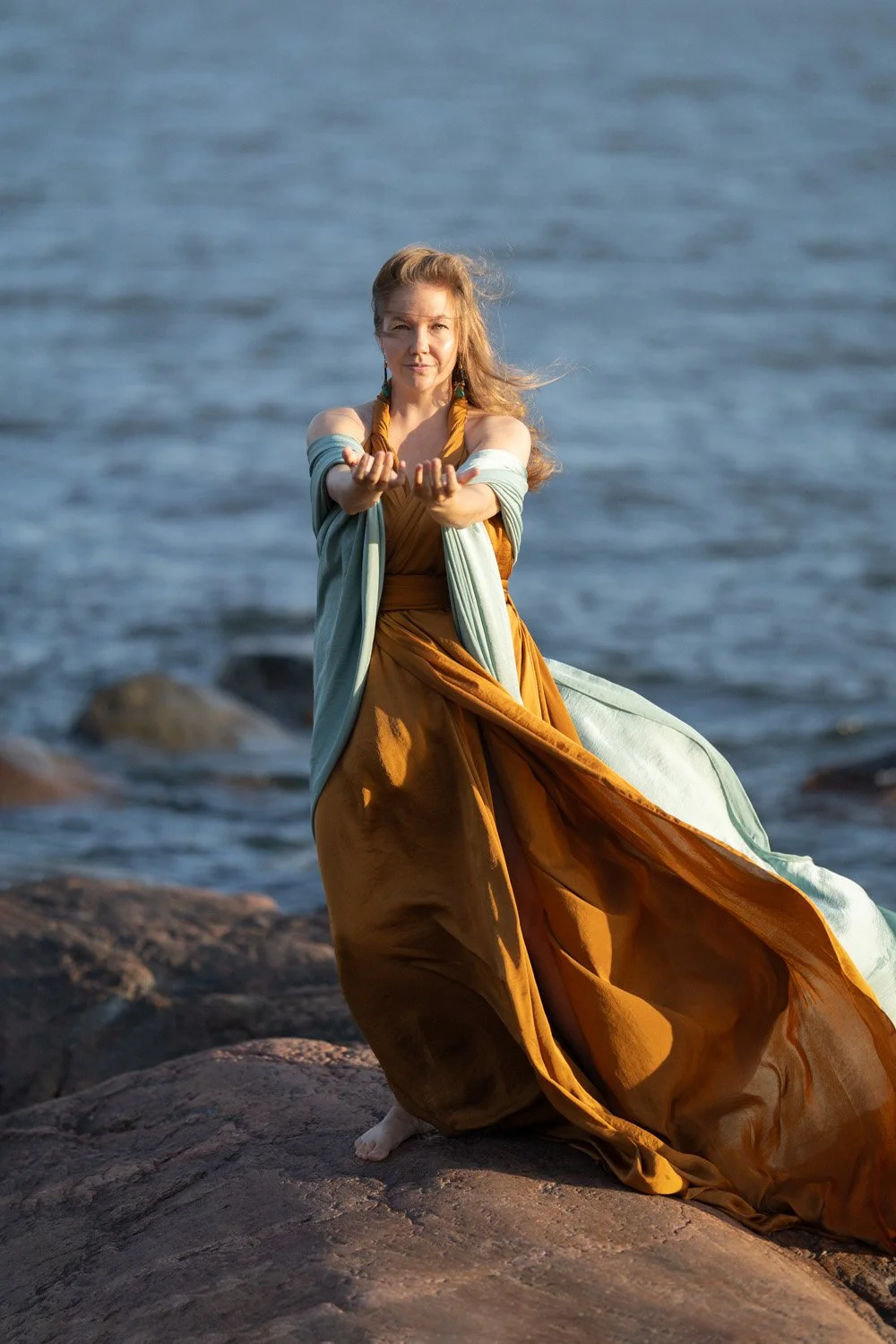 Anni Pellikka standing on a rocky shoreline in a flowing orange dress.