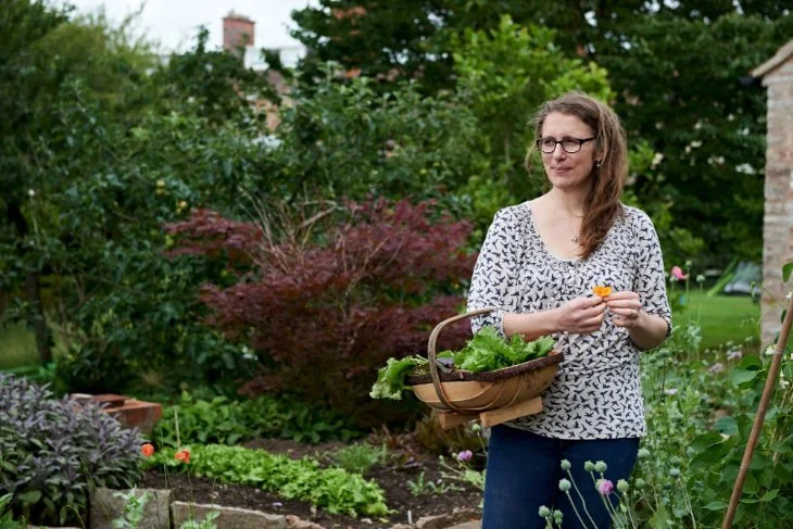 Jo Webster, nutritionist and medical herbalist, standing in her garden holding a basket of homegrown salad leaves.