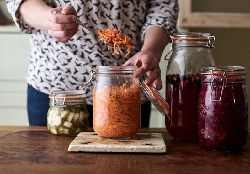Jo spooning carrot ferment from a jar, surrounded by jars of various ferments