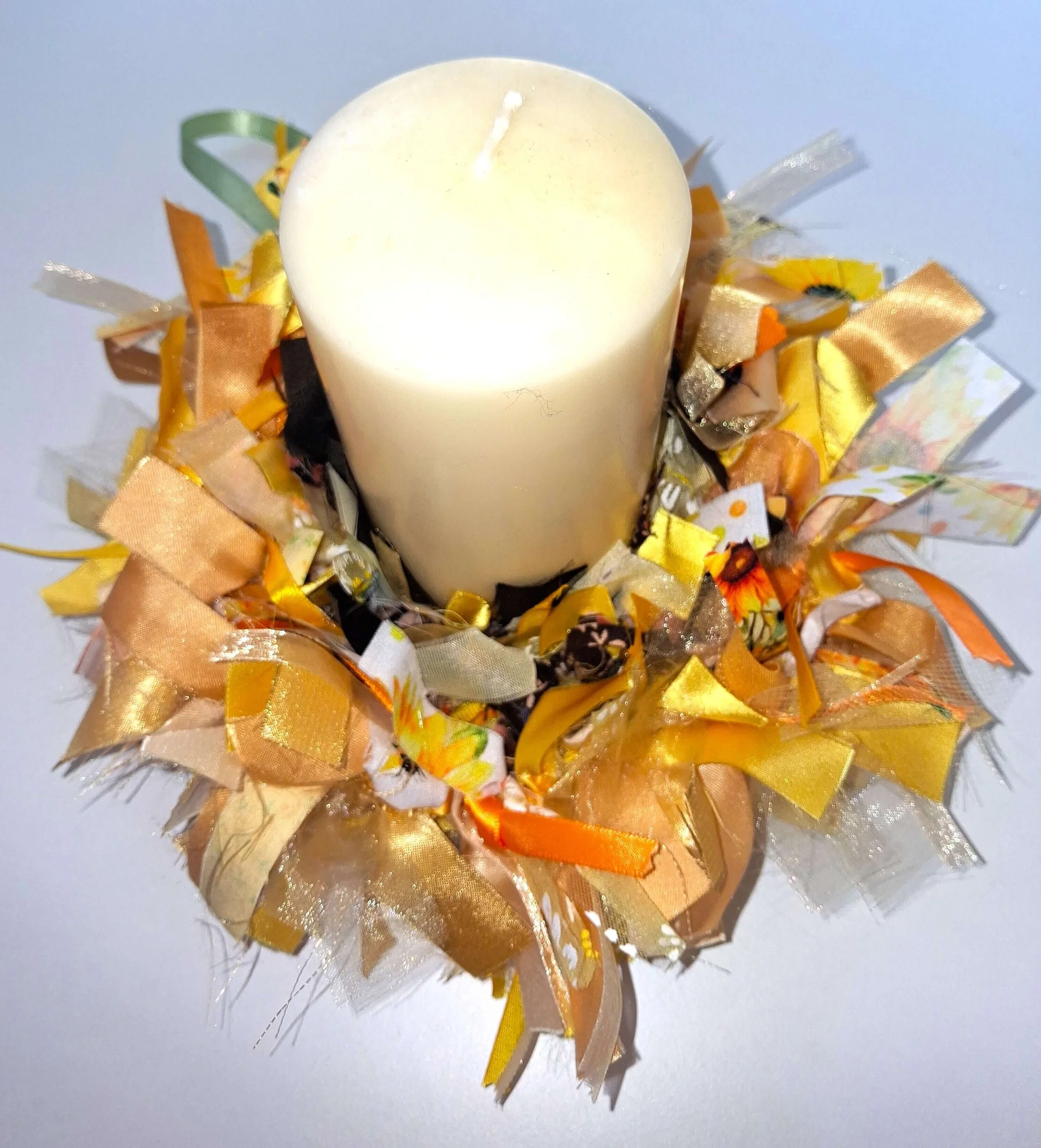 A white candle surrounded by decorative ribbons and bows in shades of gold, orange, and yellow