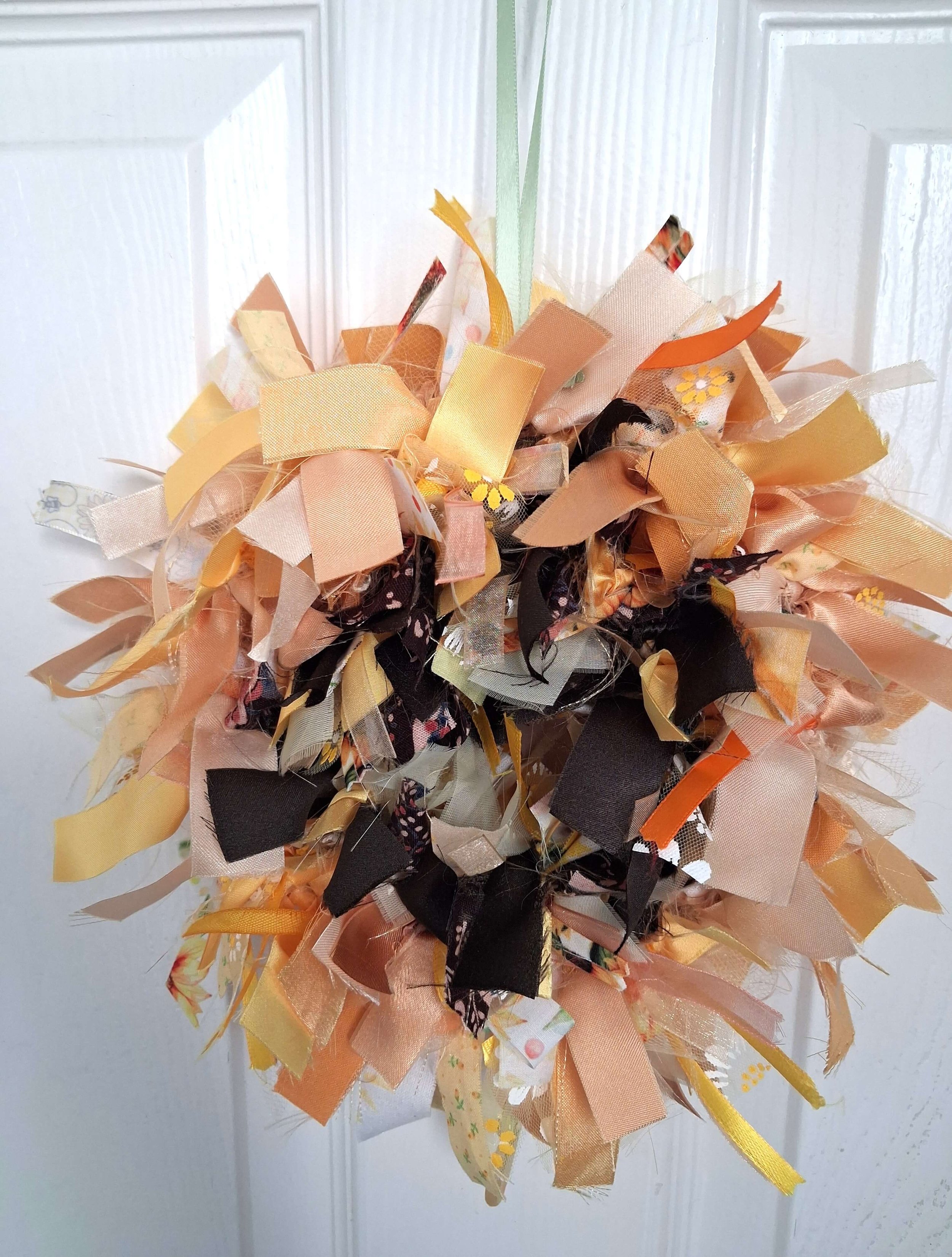 A decorative wreath made of fabric ribbons in shades of orange, peach, brown, and cream, with some floral patterns, hanging on a white door.