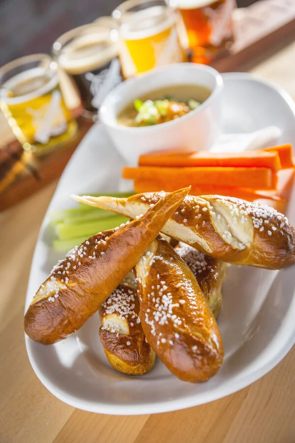 Baked pretzels sprinkled with coarse salt served with celery sticks, carrot sticks, a bowl of mustard, and three glasses of beer in the background.
