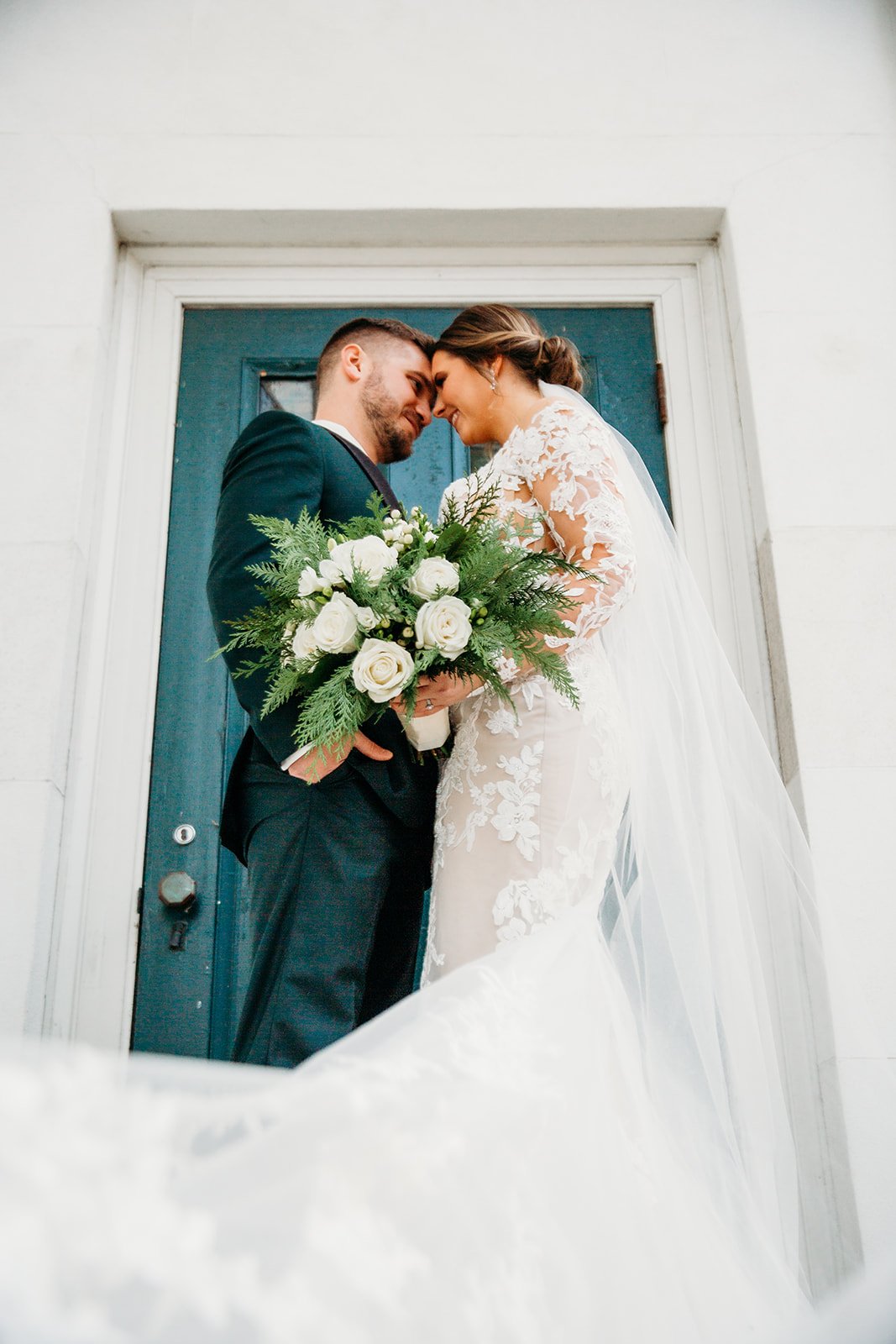 bride and groom lean heads together by teal door