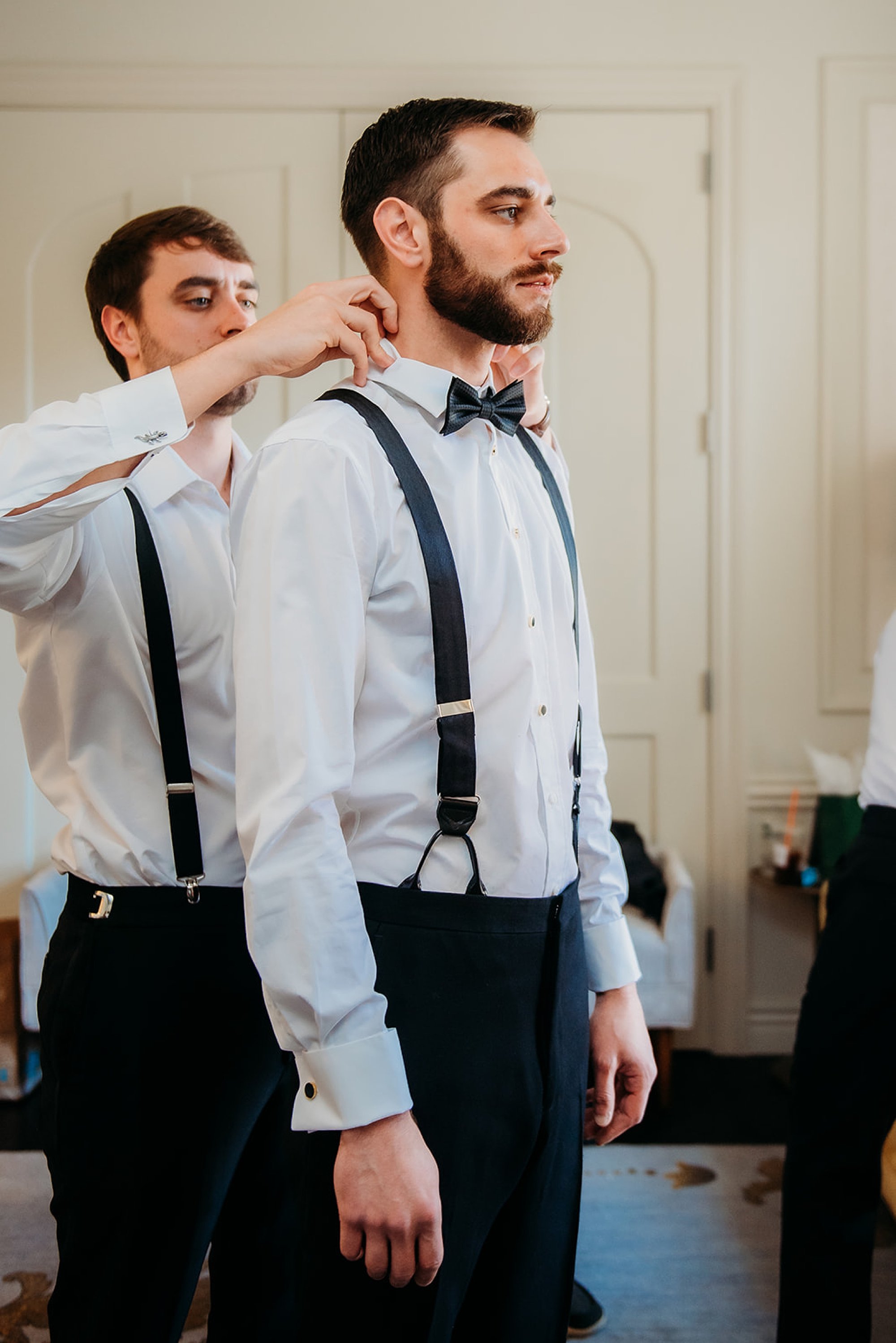 groomsman helps groom with tie and suspenders