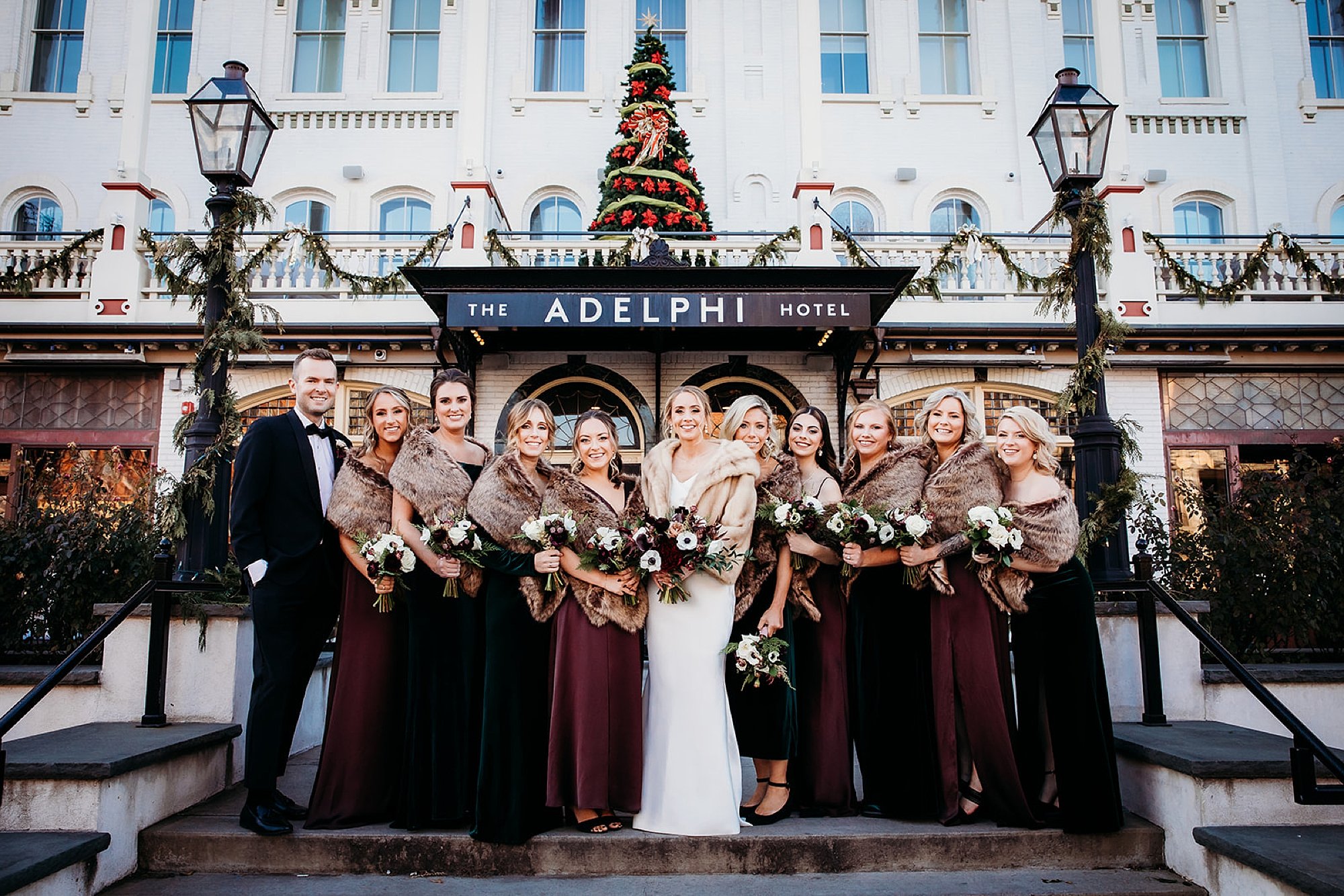 bride poses with bridesmaids and best man in burgundy gowns outside The Adelphi Hotel
