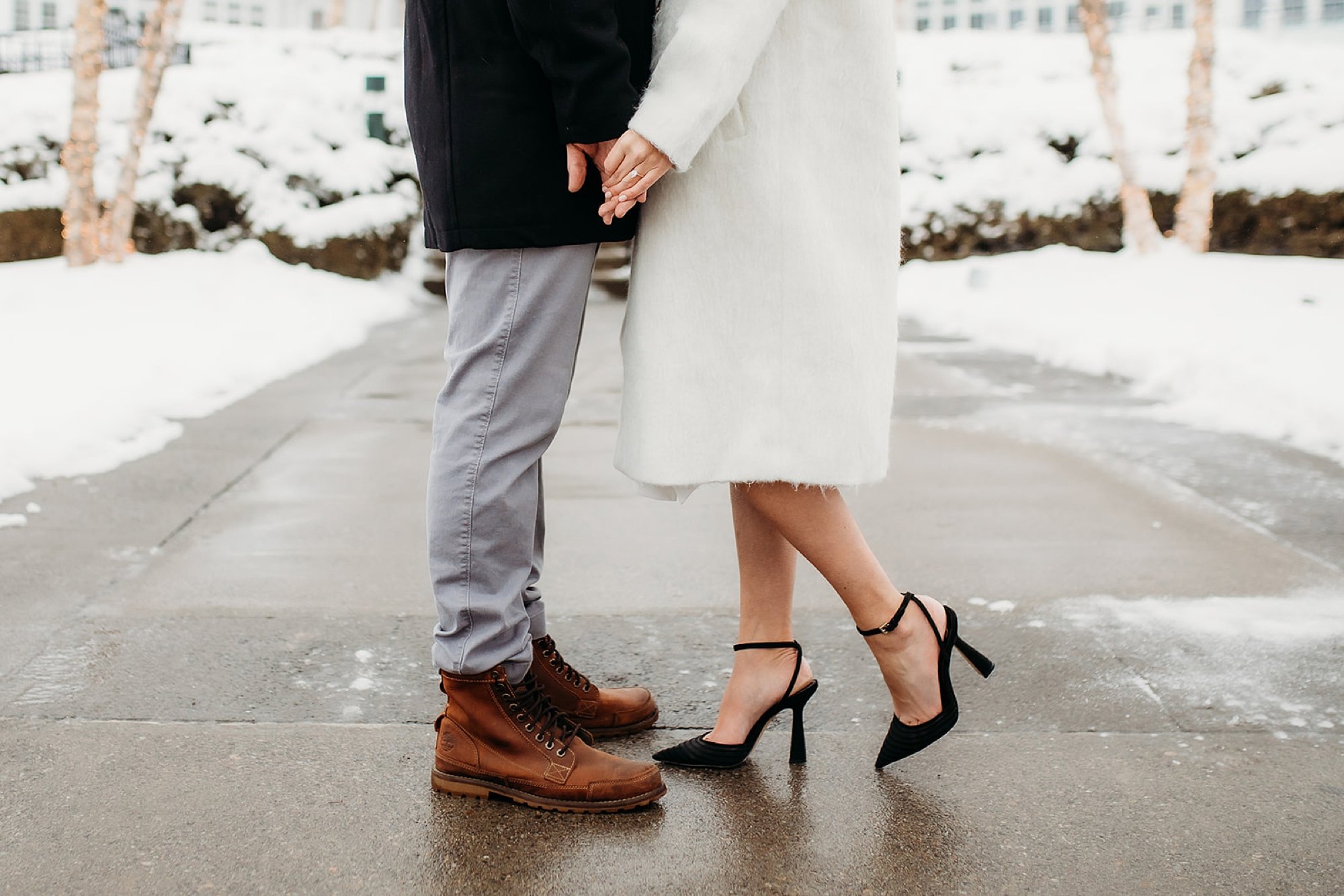 bride and groom hold hands in winter coats during snowy engagement session