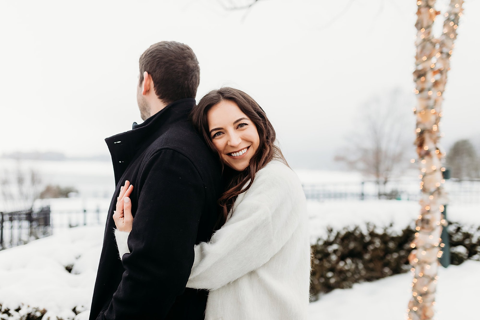 woman in white winter jacket lays head on man's back during snowy engagement session