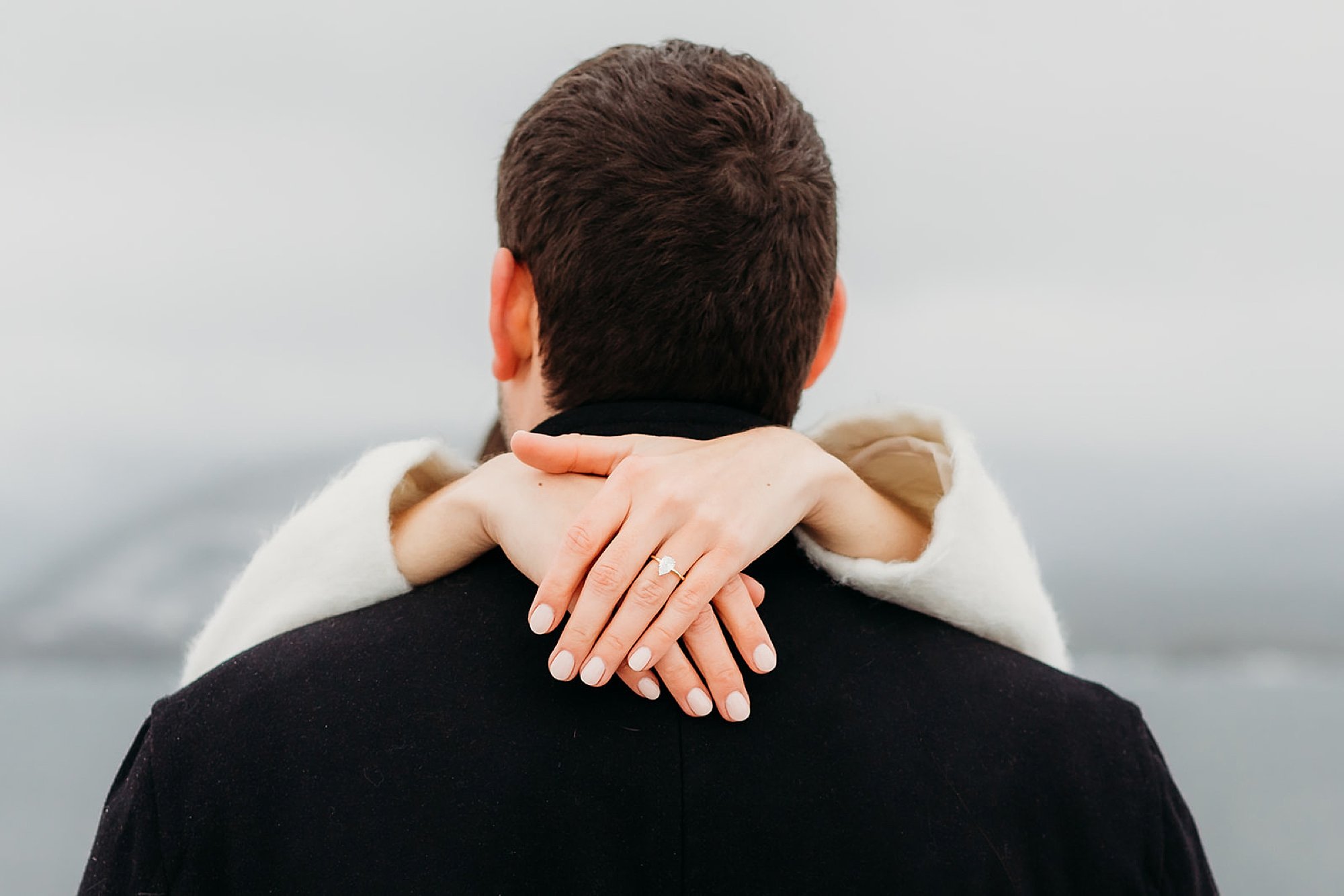 woman holds man around neck, showing off engagement ring