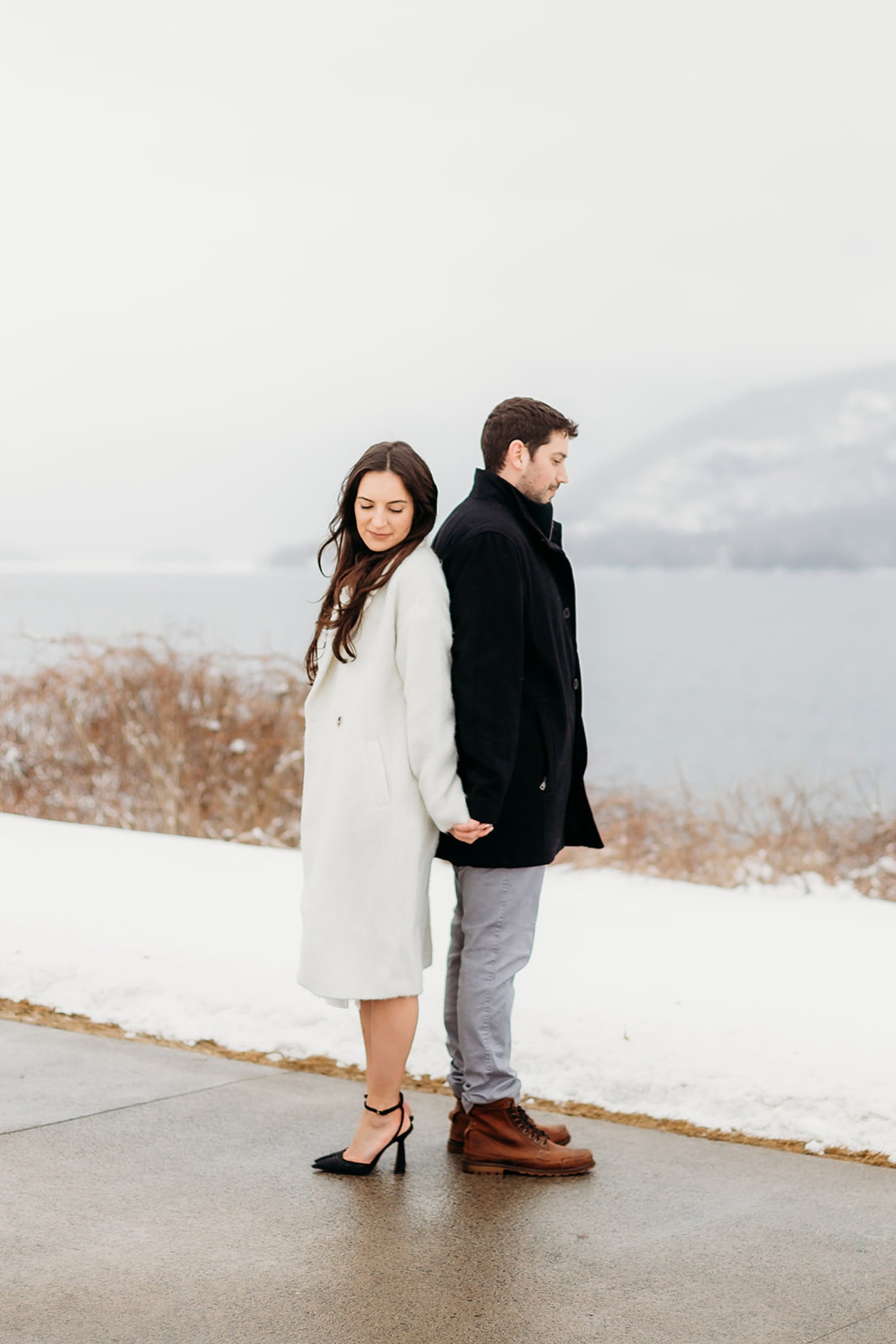 engaged couple stands back to back during winter engagement session on Lake George 