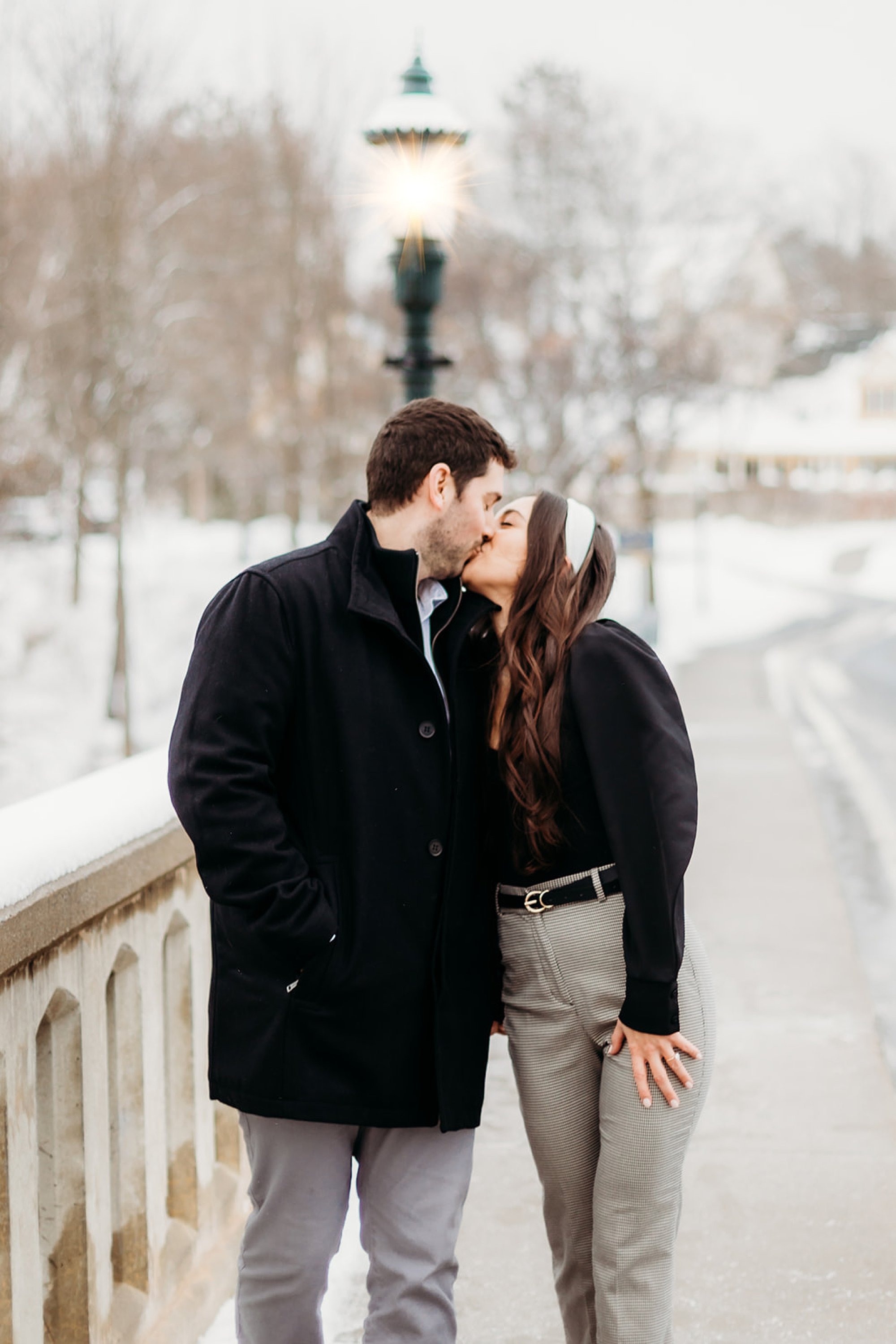 woman with brown hair kisses man in black jacket by green lamp post by Lake George