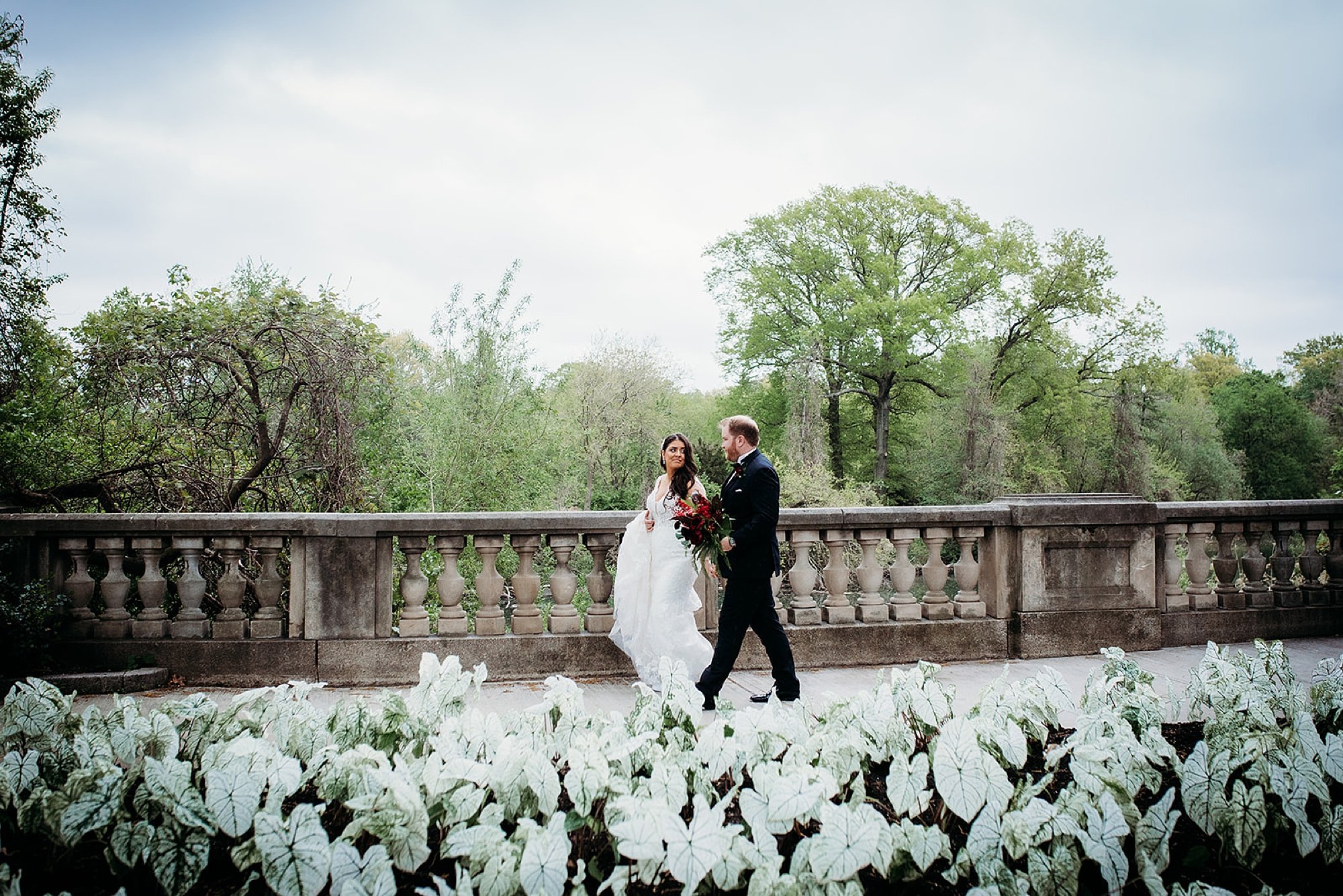 bride and groom hold hands walk on stone bridge in the Bronx Zoo