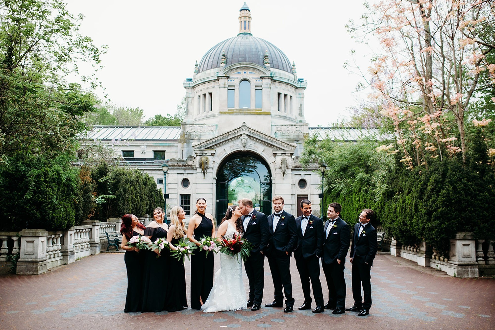 bride and groom pose with wedding party in black in front of the Bronx Zoo