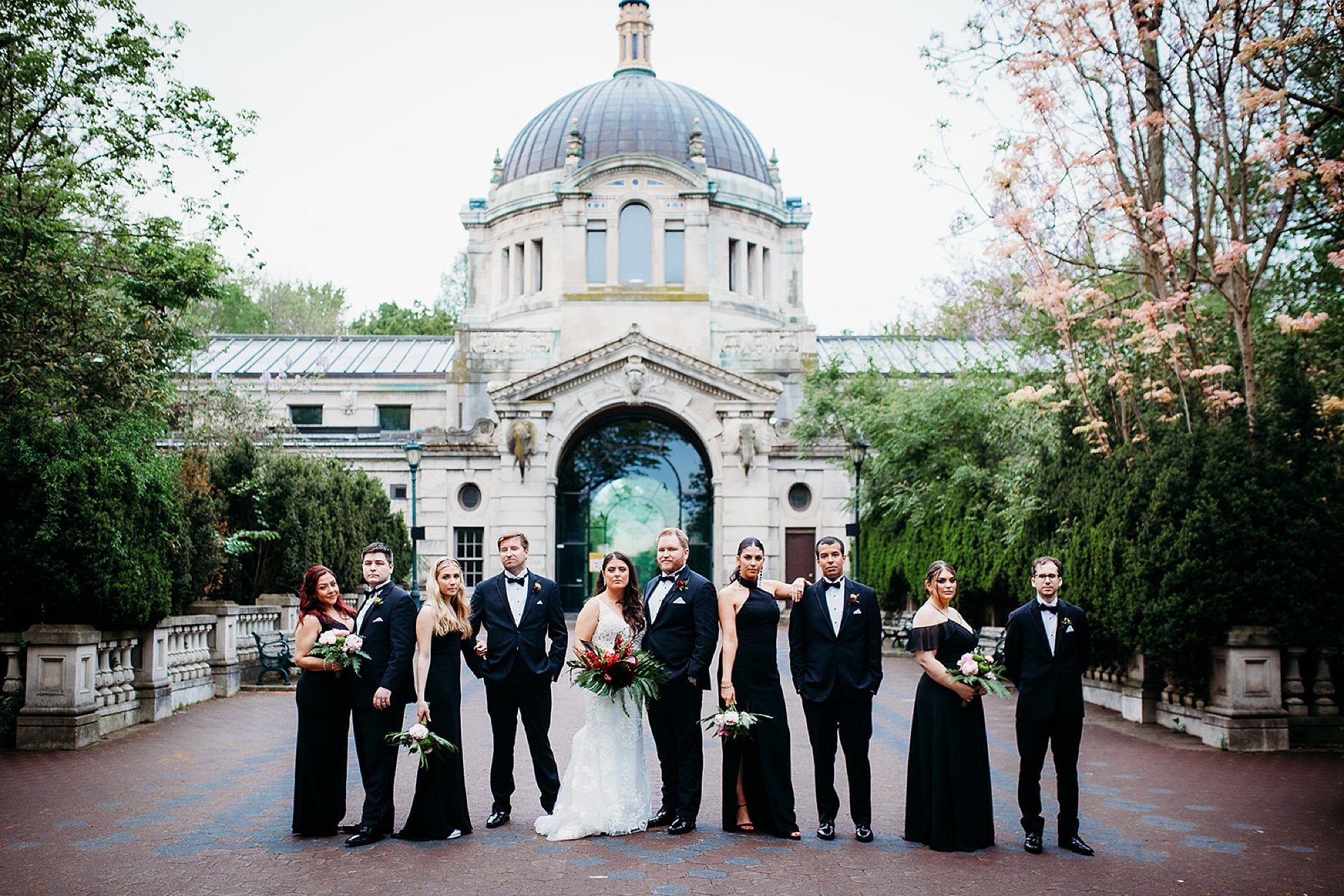 bride and groom pose with wedding party in black in front of the Bronx Zoo