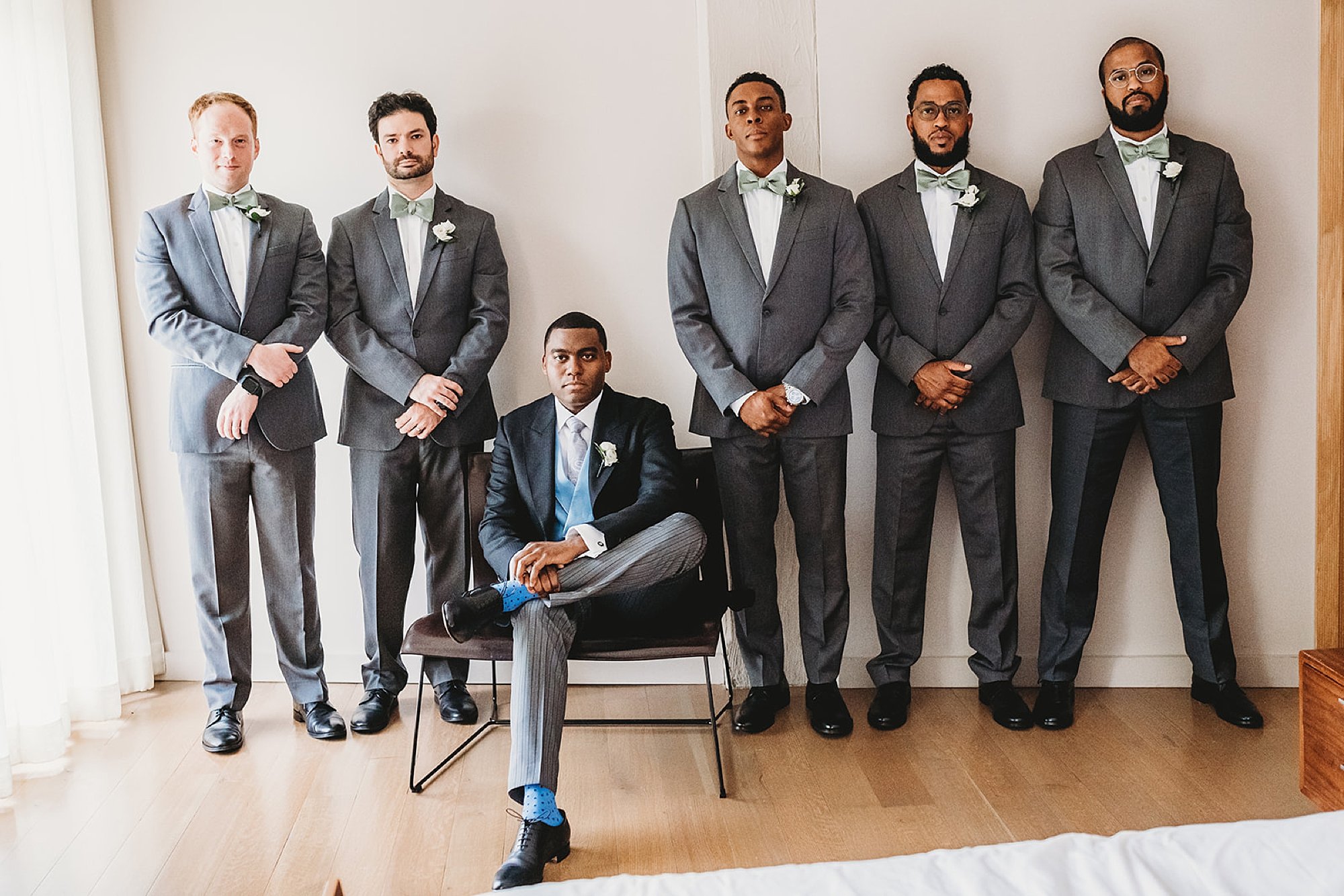 groom sits in black chair with five groomsmen standing behind him in grey suits 