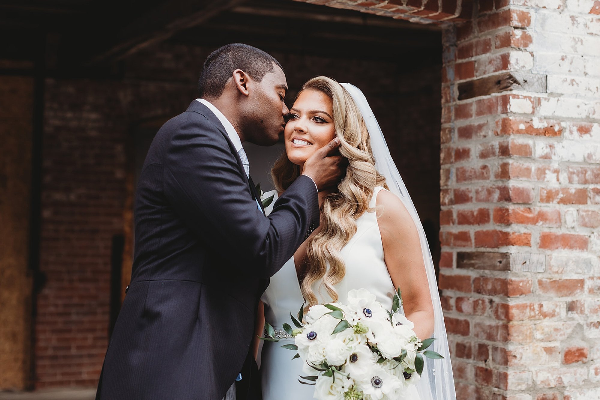 groom leans to kiss bride's cheek in front of brick building at The Roadhouse