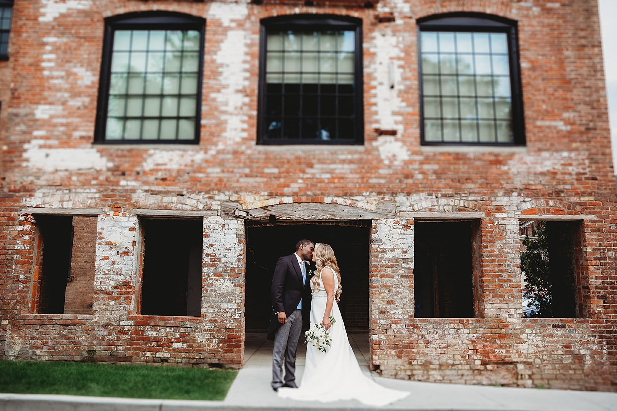 newlyweds kiss in front of brick building with glass windows at The Roadhouse
