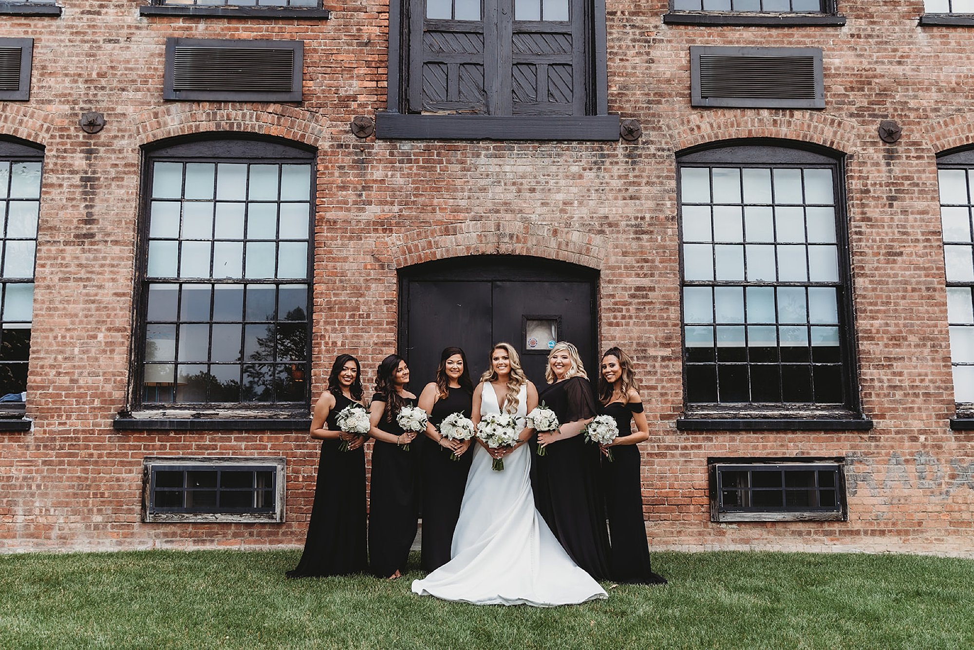 bride poses with bridesmaids in black gowns by brick building at The Roadhouse