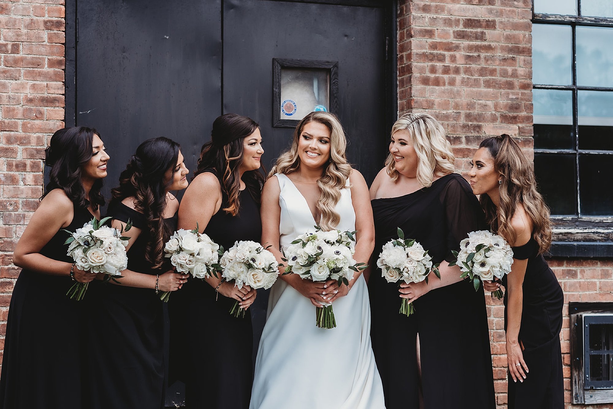 bride laughs with bridesmaids in black gowns outside brick building at The Roadhouse