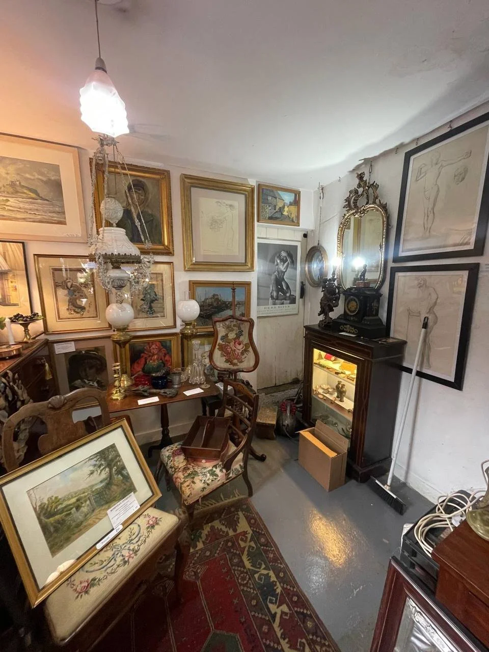 A small vintage antique shop with framed artwork and various decorative items on the walls and tables, including chairs, a glass display cabinet, and a large ornate mirror.
