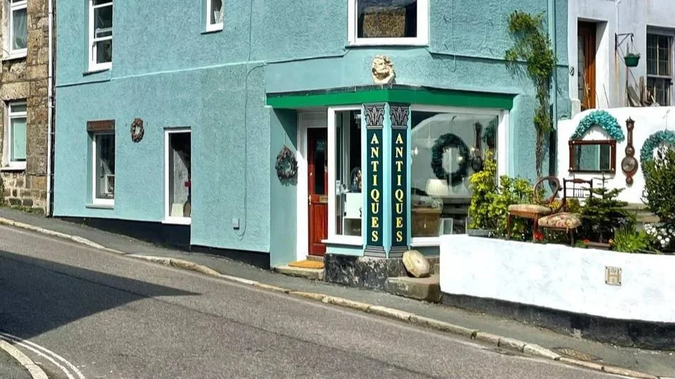 A small building with a blue facade and a green awning labeled "Antiques." The storefront has large window displays with various antiques inside. Outside, there is a garden area with plants, a decorative chair, and hanging ornaments, situated beside a white wall and next to a street.