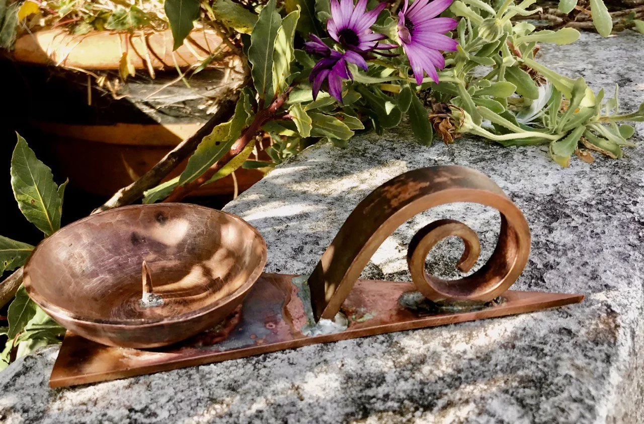 A metal sculpture resembling a spiral seashell with a shallow bowl attached, placed on a granite stone surface outdoors, with purple flowers and green leaves nearby.