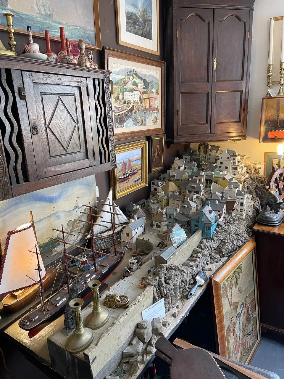 A detailed miniature model of a coastal village with houses, a bridge, and boats, displayed on a table with surrounding artworks and collectibles in a room.