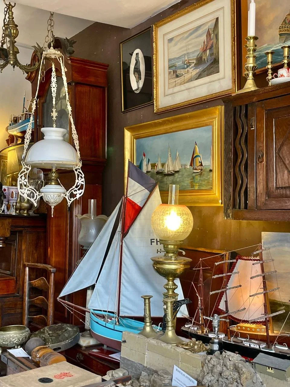 A collection of nautical and vintage decorative items arranged in a room, including model sailboats, framed landscape and maritime paintings, an ornate lamp, and various antique objects.