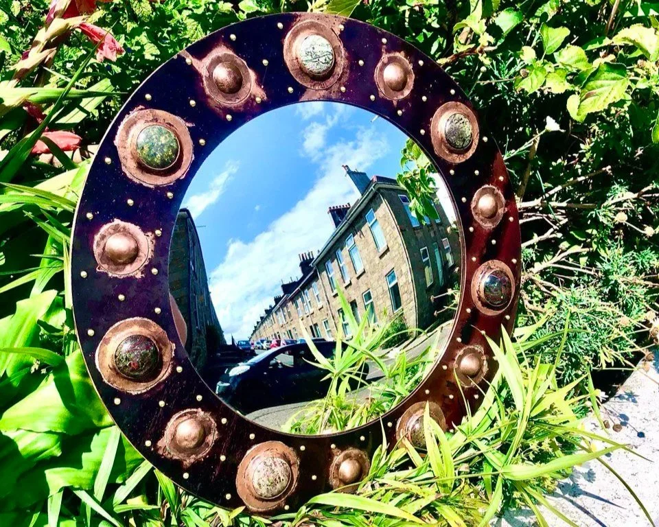 A round decorative mirror with a vintage-style copper frame surrounded by green plants, reflecting a street scene with buildings and a blue sky.